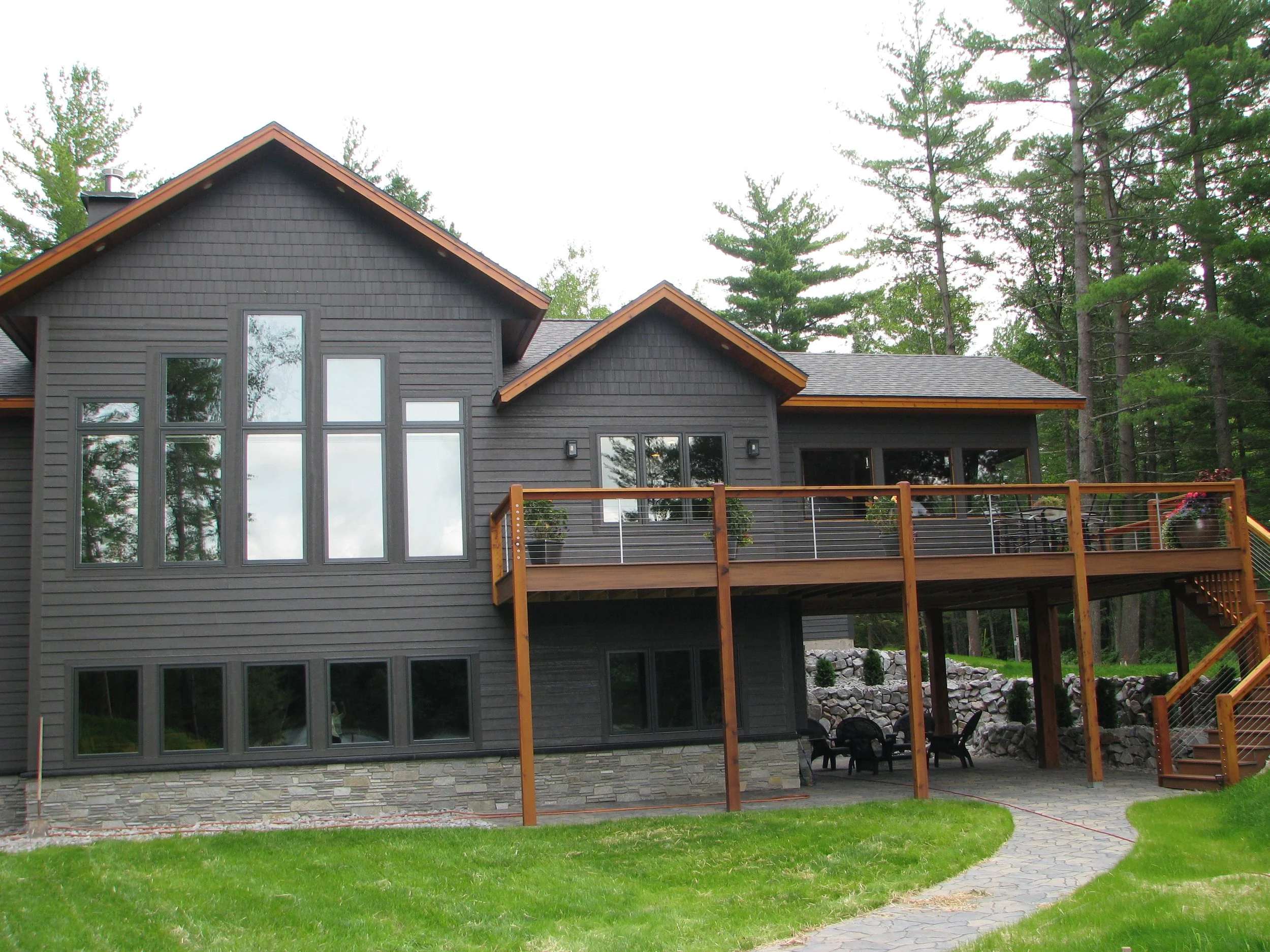 A modern, dark gray house with large windows, a wooden deck, and outdoor seating, surrounded by trees and a grassy lawn.