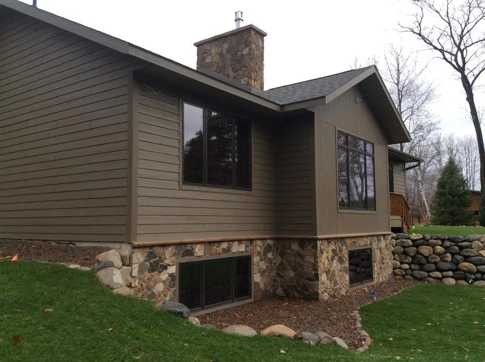 Side view of a house with stone foundation, beige siding, multiple windows, a chimney, and a small deck with wooden railing, surrounded by trees and a grassy yard.