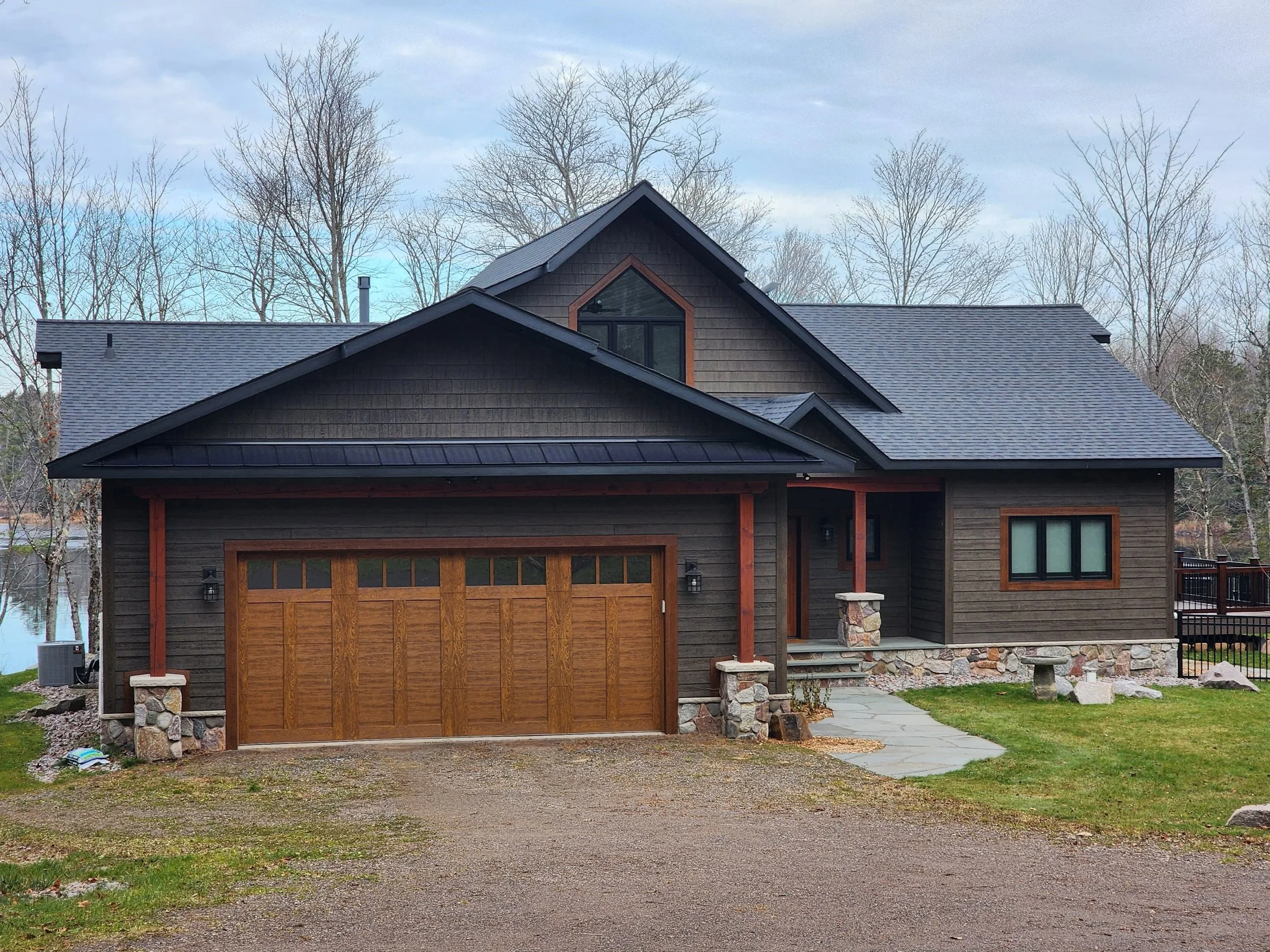 A modern two-story house with dark brown wooden siding, a black metal roof, a wooden garage door, stone accents at the base, a small front porch with stone stairs, and surrounded by a grassy lawn with leafless trees in the background.