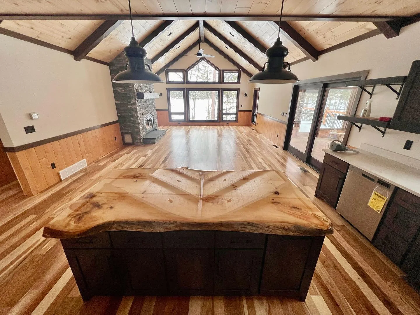 Empty open-concept living room with vaulted wood ceiling, large windows, and a stone fireplace. A kitchen island with a wooden countertop is in the foreground, and sliding glass doors lead to an outdoor deck.