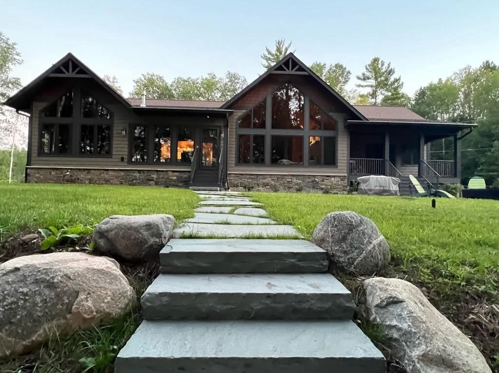A modern house with large glass windows, a stone foundation, and a wooden exterior surrounded by a grassy yard with rocks and a paved stone walkway leading to the front door.