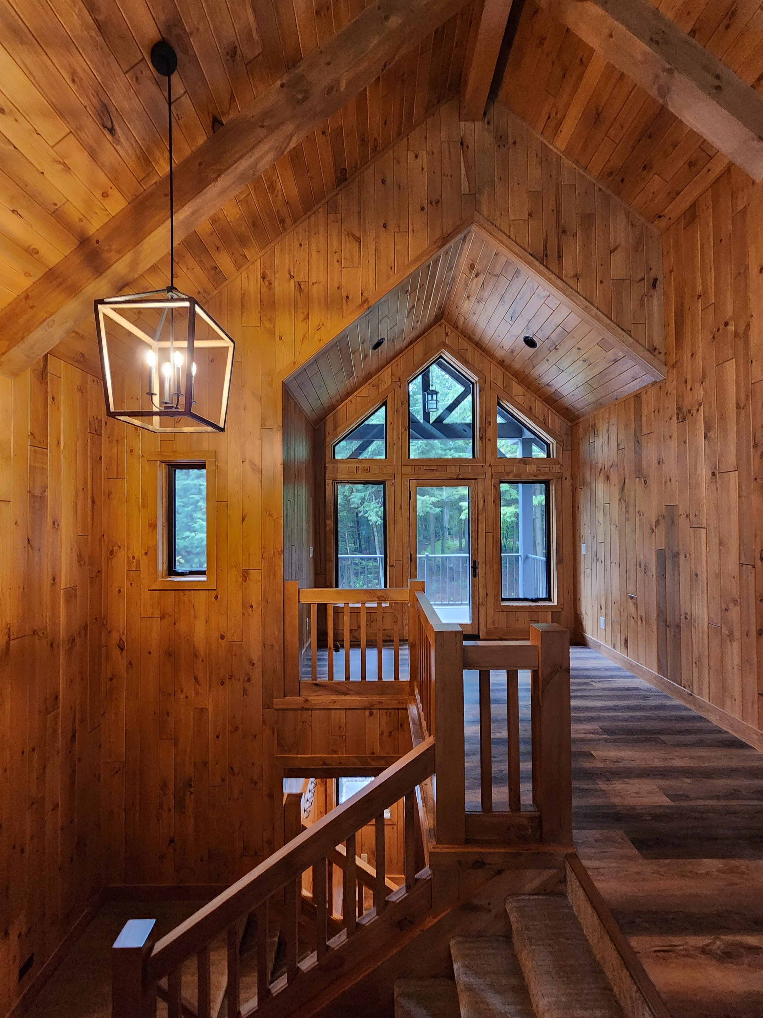Wood-paneled interior of a cabin or house with a vaulted ceiling, large triangular window, staircase, and a modern pendant light.