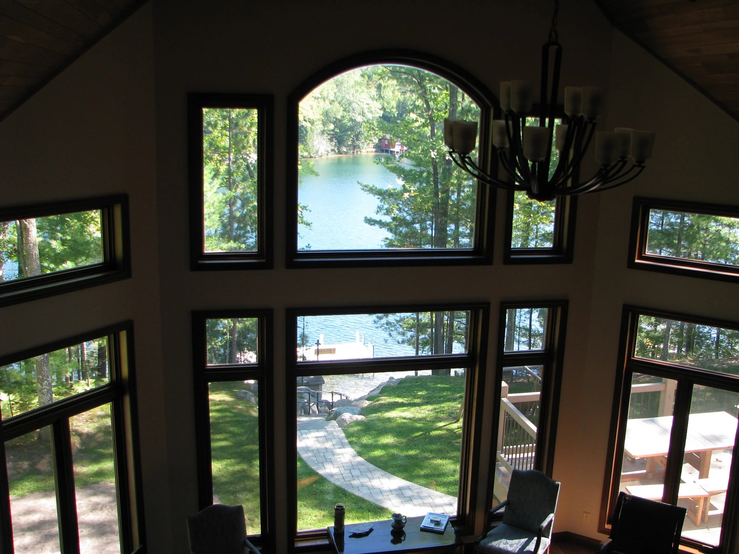 Interior view of a house looking out through large windows at a lake with trees and a dock, with a chandelier hanging from the ceiling.