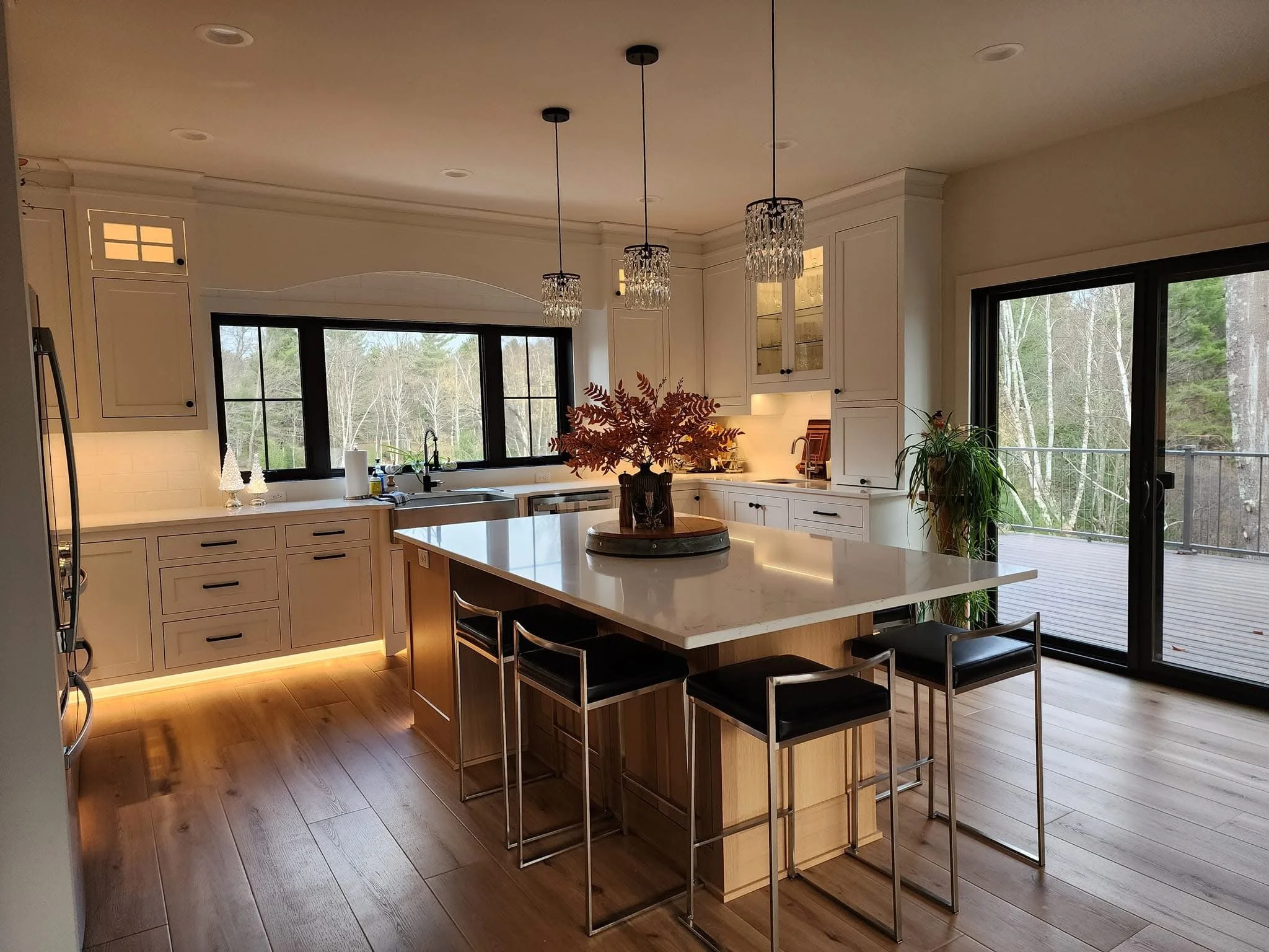 Modern kitchen with white cabinets, a large island with a marble countertop, black bar stools, pendant lights, large windows showing outdoor scenery, and a sliding glass door leading to a deck.