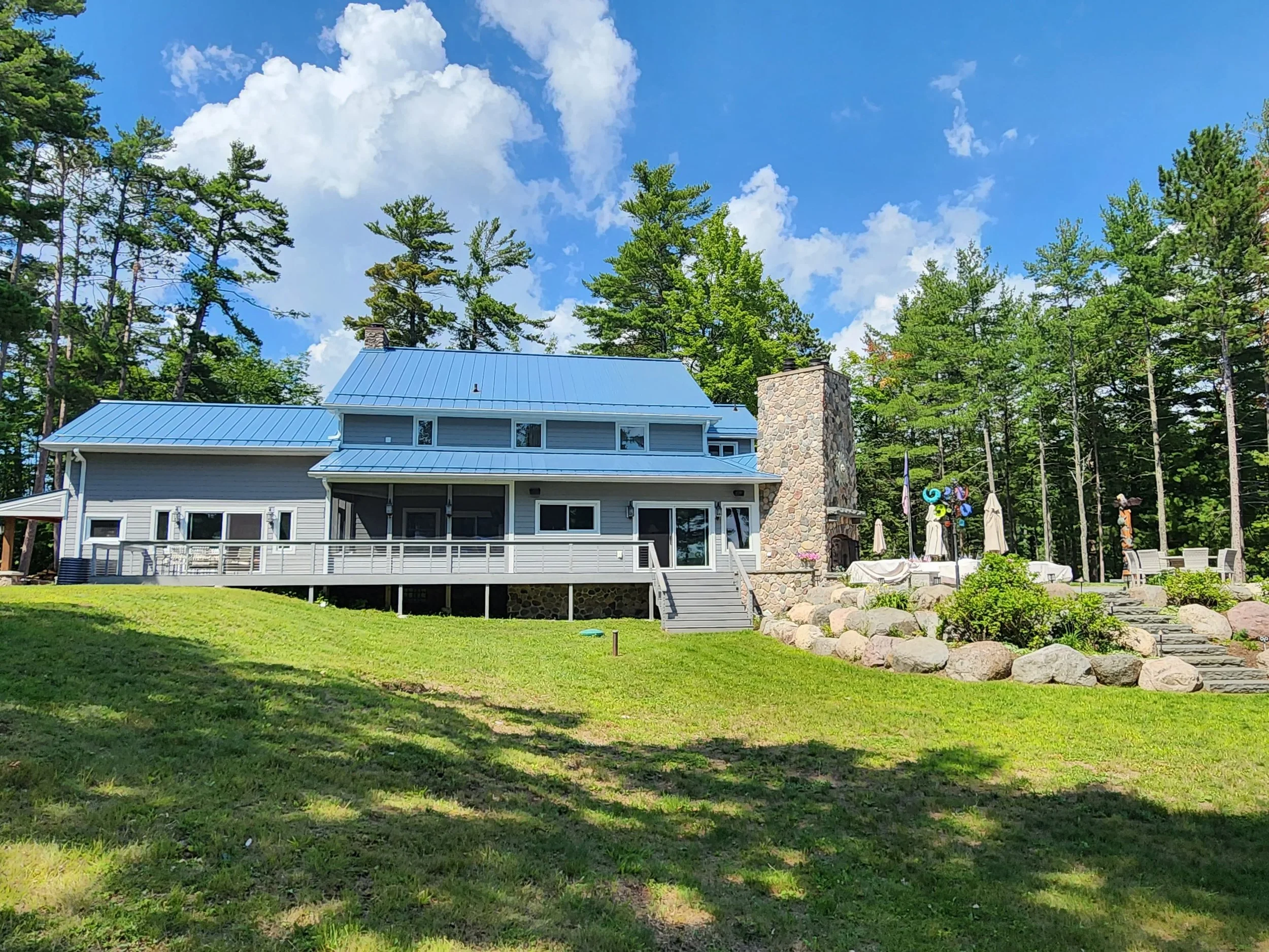 A two-story house with a blue metal roof and a large stone chimney, surrounded by trees and a well-maintained green lawn, with a backyard patio area with umbrellas and colorful wind spirals.