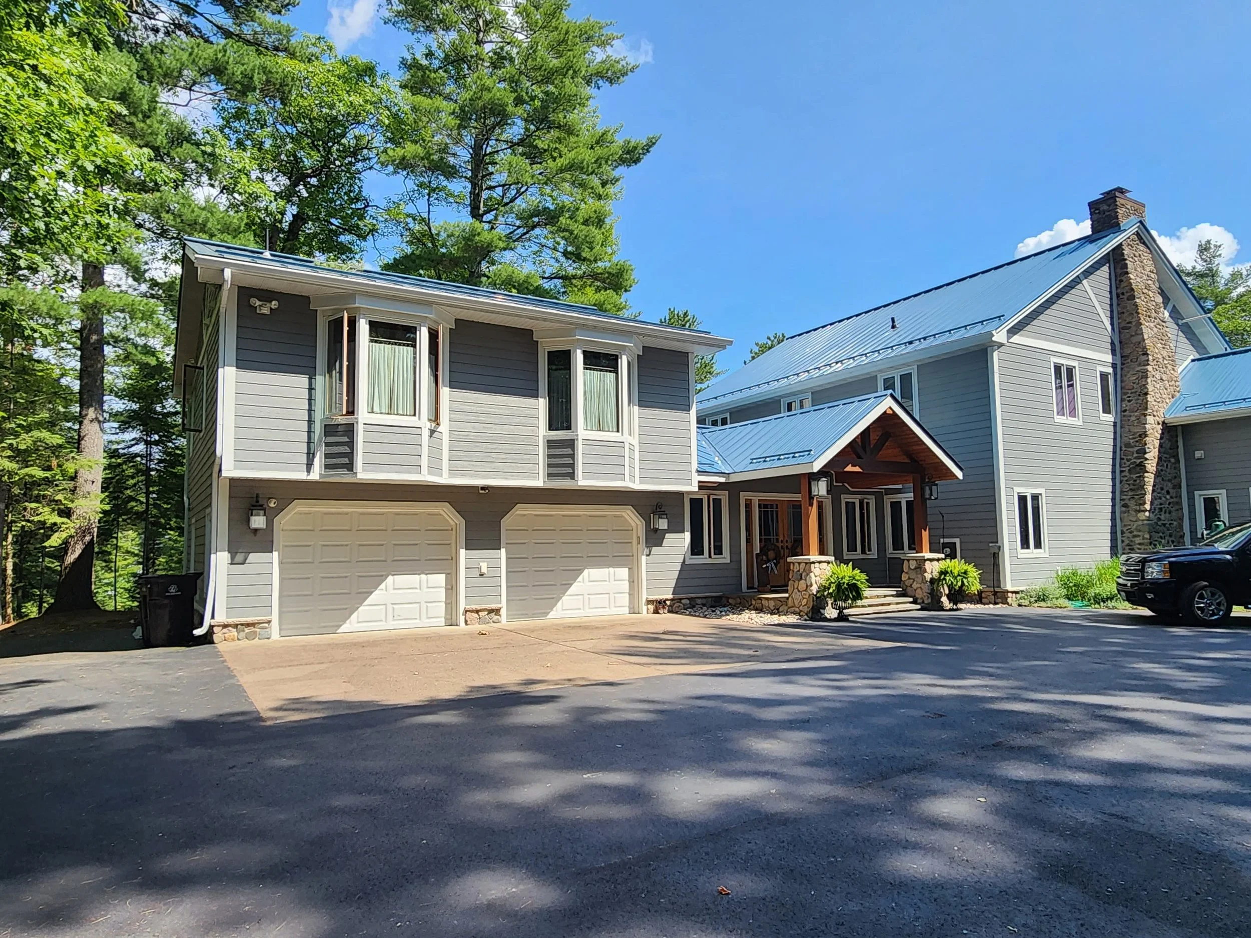 A modern house with gray siding, a metal roof, and a stone chimney, surrounded by trees and a paved driveway.
