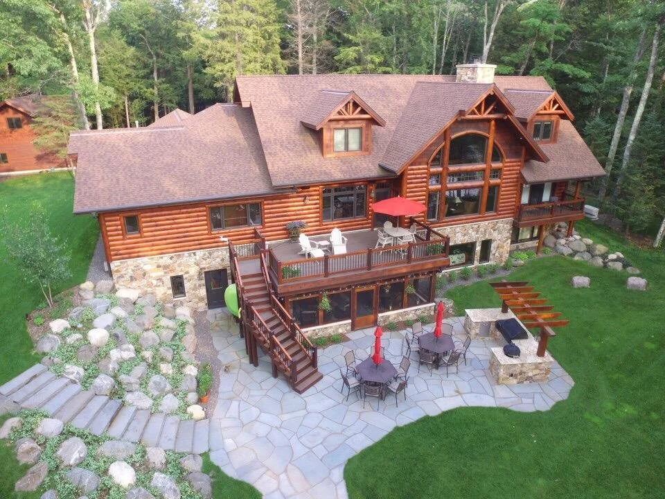 Large log cabin-style house with a stone foundation, multiple decks with outdoor furniture, and an outdoor fireplace, surrounded by trees and greenery.