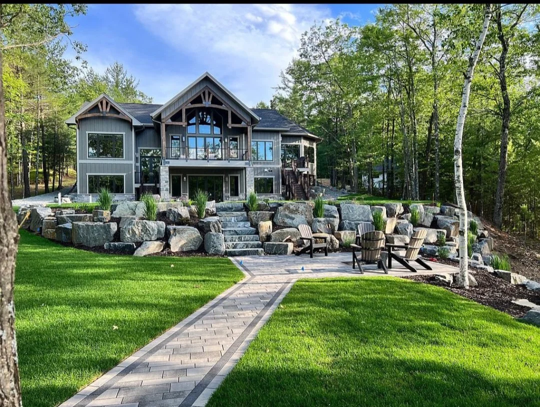 A modern two-story house with large windows, surrounded by a landscaped yard with a stone retaining wall, stone steps, green grass, and outdoor seating on a patio. Trees and a forested area are in the background.