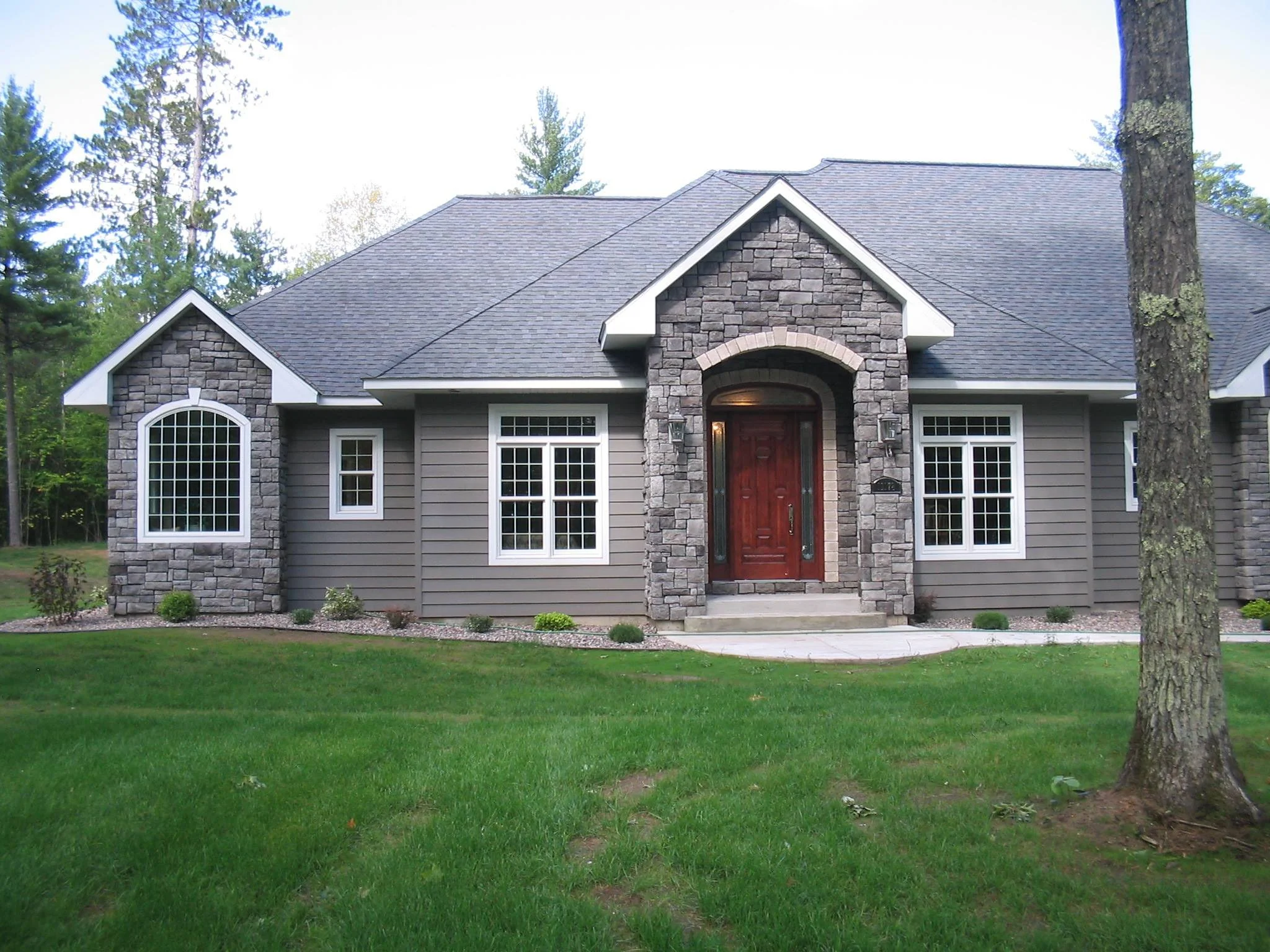 A house with gray siding, stone accents, and a gabled roof. It has multiple windows with white trim and a wooden front door. The house is surrounded by a green lawn and trees.