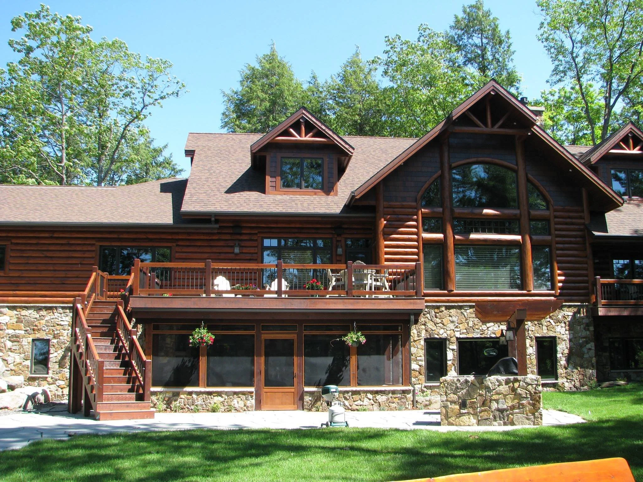 A large wooden house with stone foundation, multiple windows, a deck with chairs, and a staircase leading to the backyard, surrounded by green trees and grass.