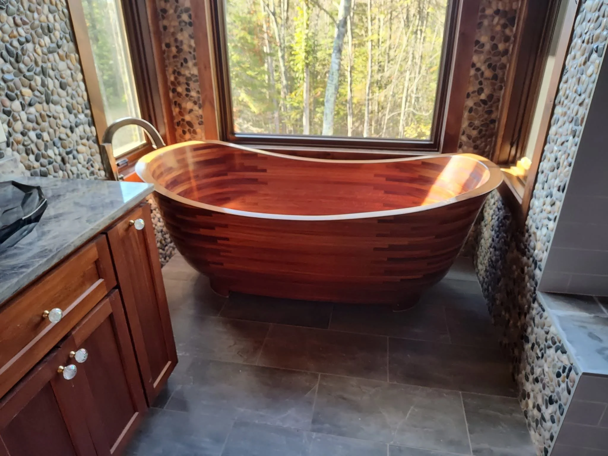 Wooden bathtub near a large window in a bathroom with pebble walls and cabinetry with marble countertop.
