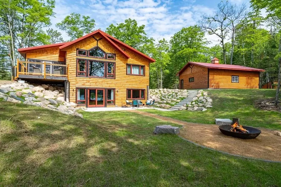 Two log cabins in a wooded area with a grassy yard, a fire pit, and stone steps.