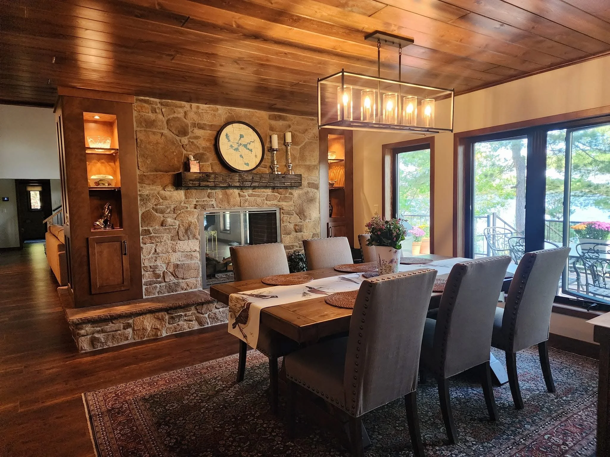 Dining room with a wooden dining table and eight upholstered chairs, a stone fireplace with a mantel, a clock and candlesticks, large windows overlooking a balcony with flowers, and a modern chandelier hanging from a wooden ceiling.
