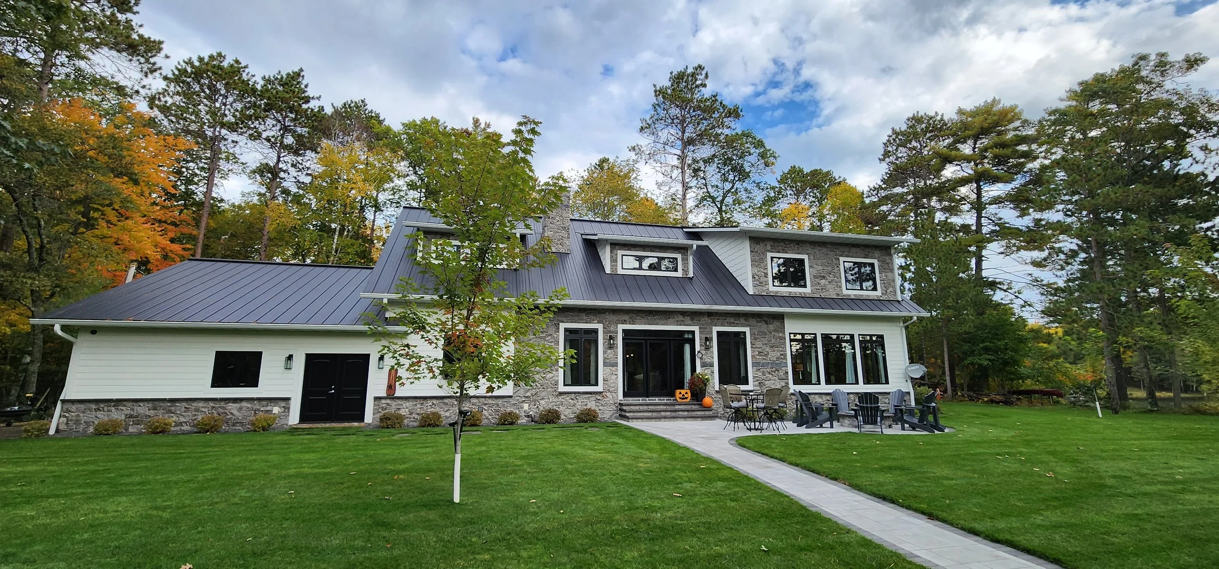 A modern two-story house with gray stone and white siding exterior, black roof, and large windows, surrounded by a well-kept green lawn and some trees with autumn leaves. There is a pathway leading to the front porch with outdoor furniture and decora