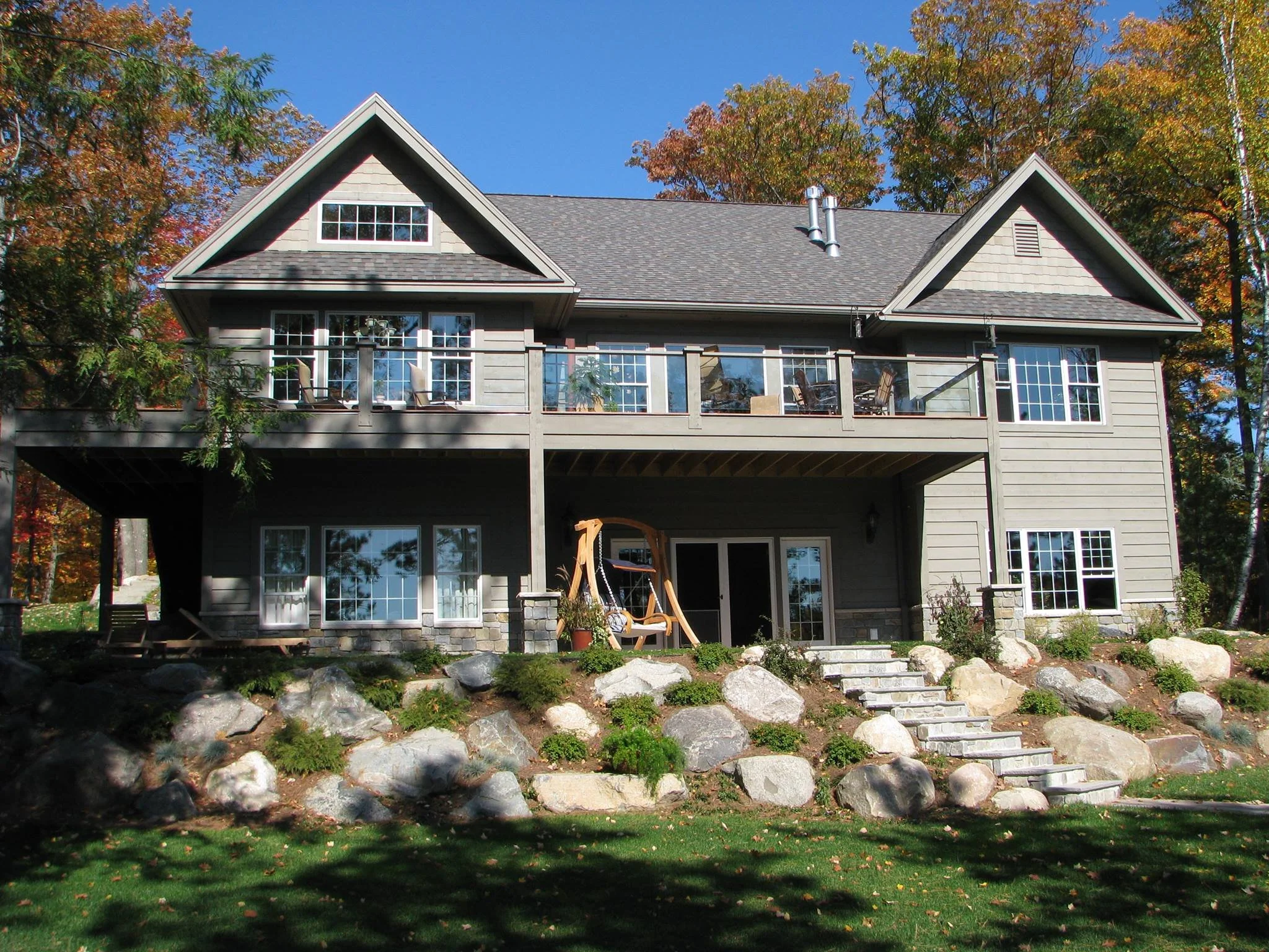 A multi-story house with gray siding, a shingled roof, and a large balcony on the upper level with patio furniture. The house is surrounded by trees with fall foliage and has large rocks and stone steps leading up to it from a grassy yard.