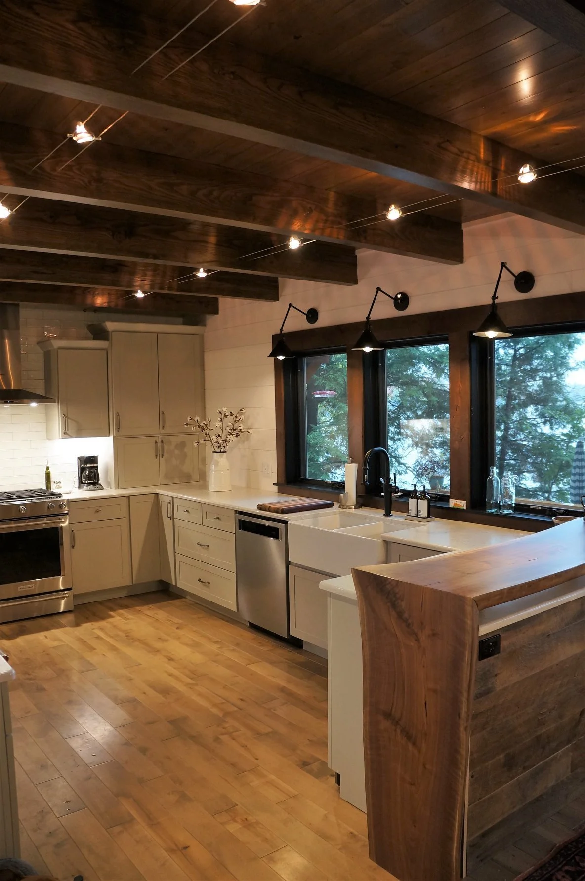 Kitchen with wood ceiling and flooring, white cabinets, black window frames, farmhouse sink, and pendant lights.
