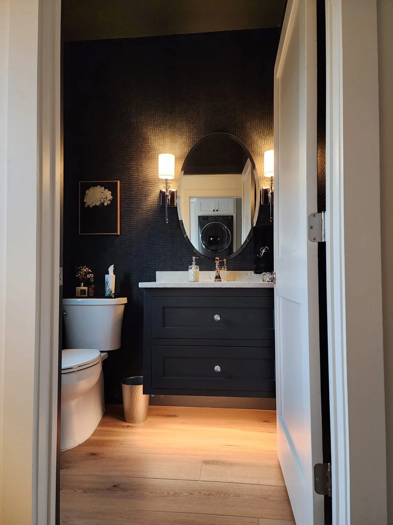 View of a modern bathroom with a black vanity, oval mirror, black textured wall, white countertop, and wall-mounted lights. A washing machine is visible in the reflection, and the bathroom has wooden flooring.
