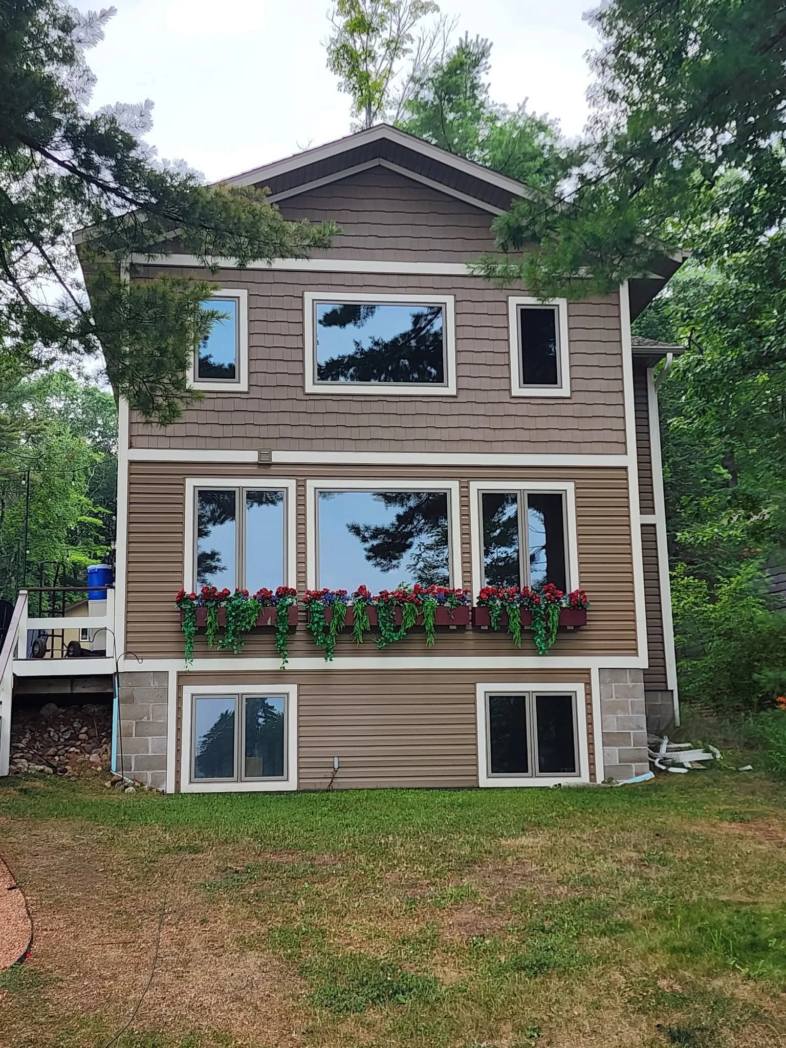 Front view of a three-story house with brown siding, white trim, and large windows, some with flower boxes filled with red and pink flowers, surrounded by green trees.