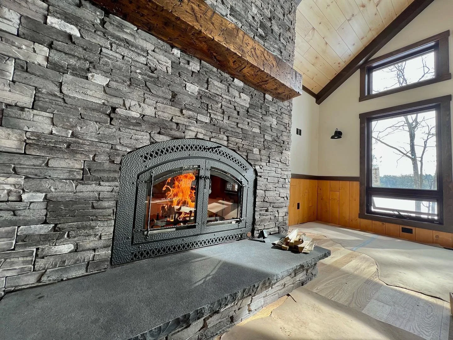 A cozy living room corner with a stone fireplace and a wood mantel. The fireplace has a burning fire inside with flames visible through a glass door. The room features large windows with dark trim, allowing natural light to fill the space. The walls are partly wood-paneled, and the ceiling is vaulted with exposed wood beams.