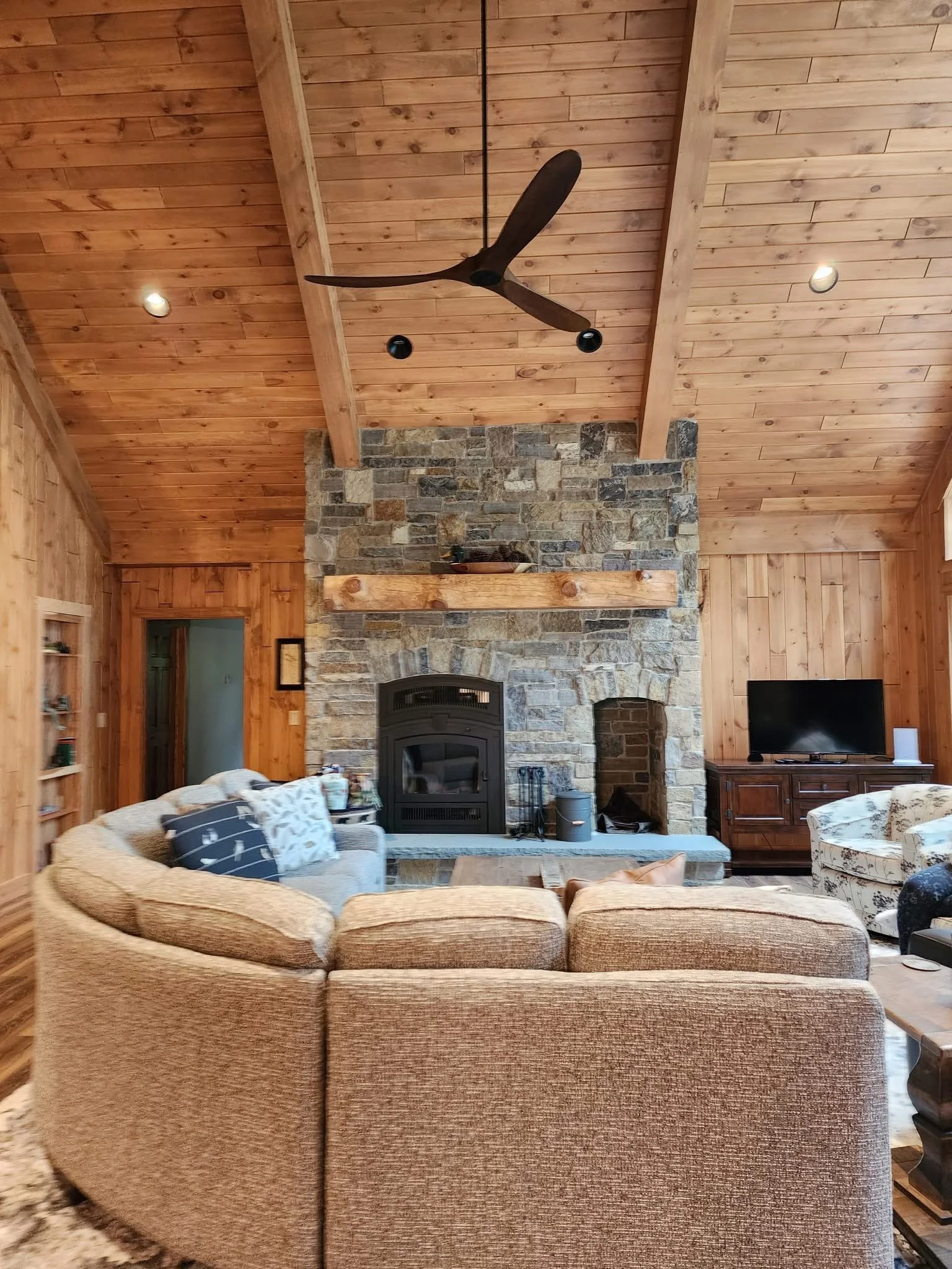 Living room with a wood-paneled ceiling, stone fireplace, ceiling fan, and various seating including a rounded beige sofa and a floral armchair. A television is on a wooden stand on the right side.
