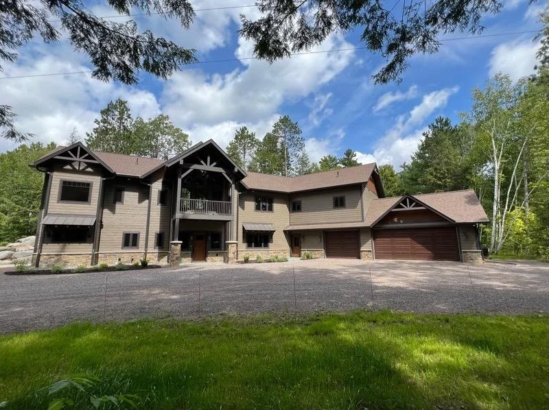 A large multi-story house with a brown exterior surrounded by trees and a gravel driveway.