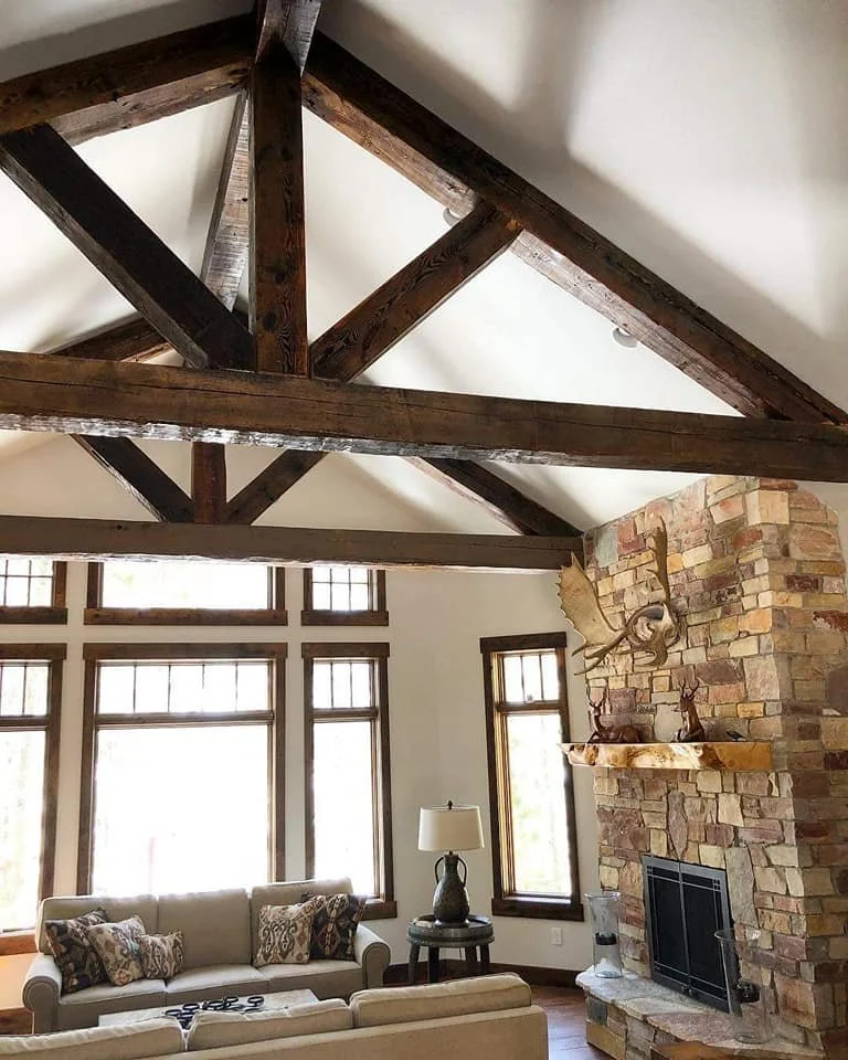 Living room with high vaulted ceiling and large windows, a stone fireplace with a wooden mantle, a sofa with patterned pillows, a side table with a lamp, and rustic wooden beams on the ceiling.
