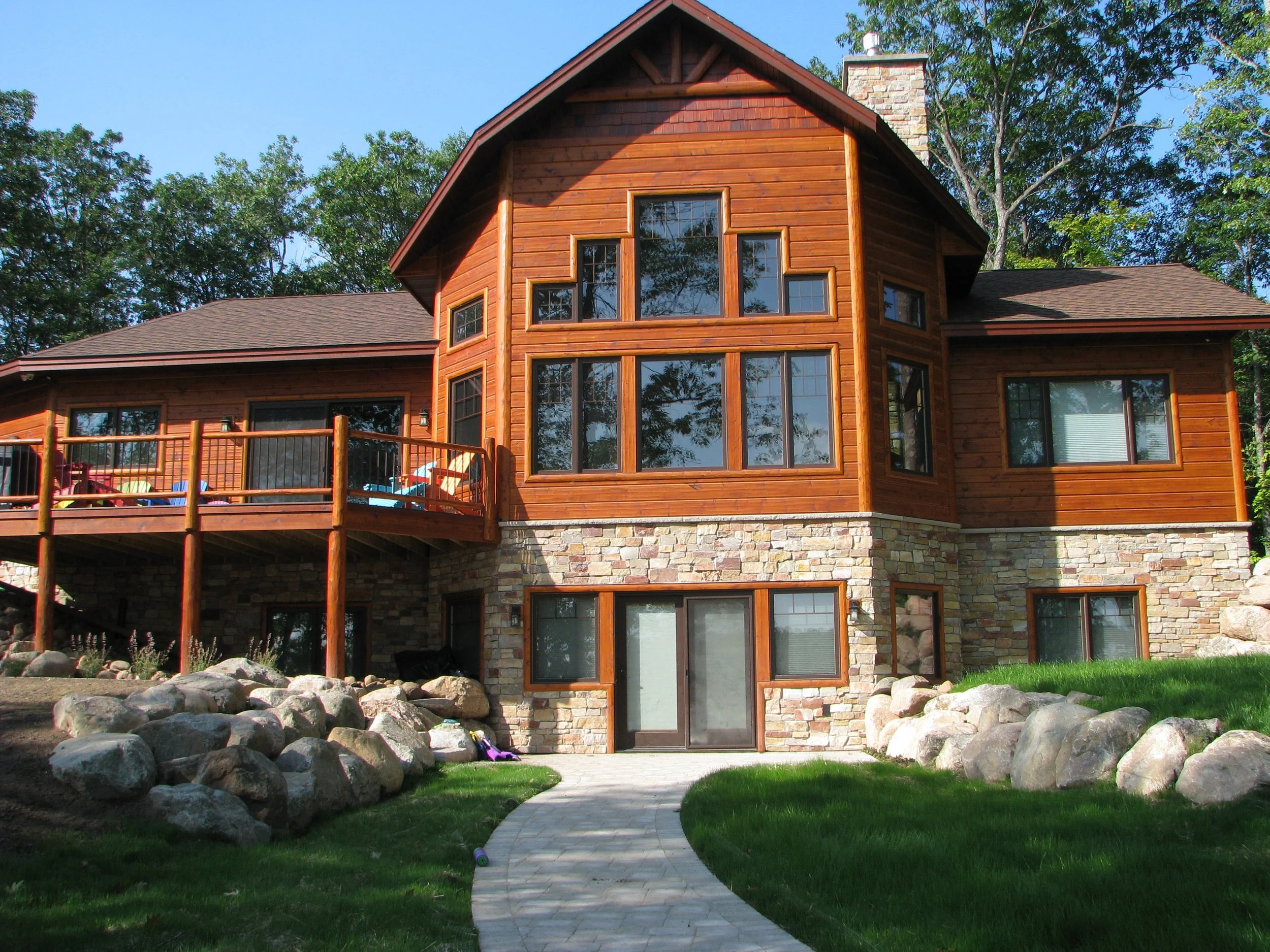 A multi-story house with a combination of wood and stone exterior, large windows, a balcony, and a paved walkway leading to glass sliding doors surrounded by green grass and trees.