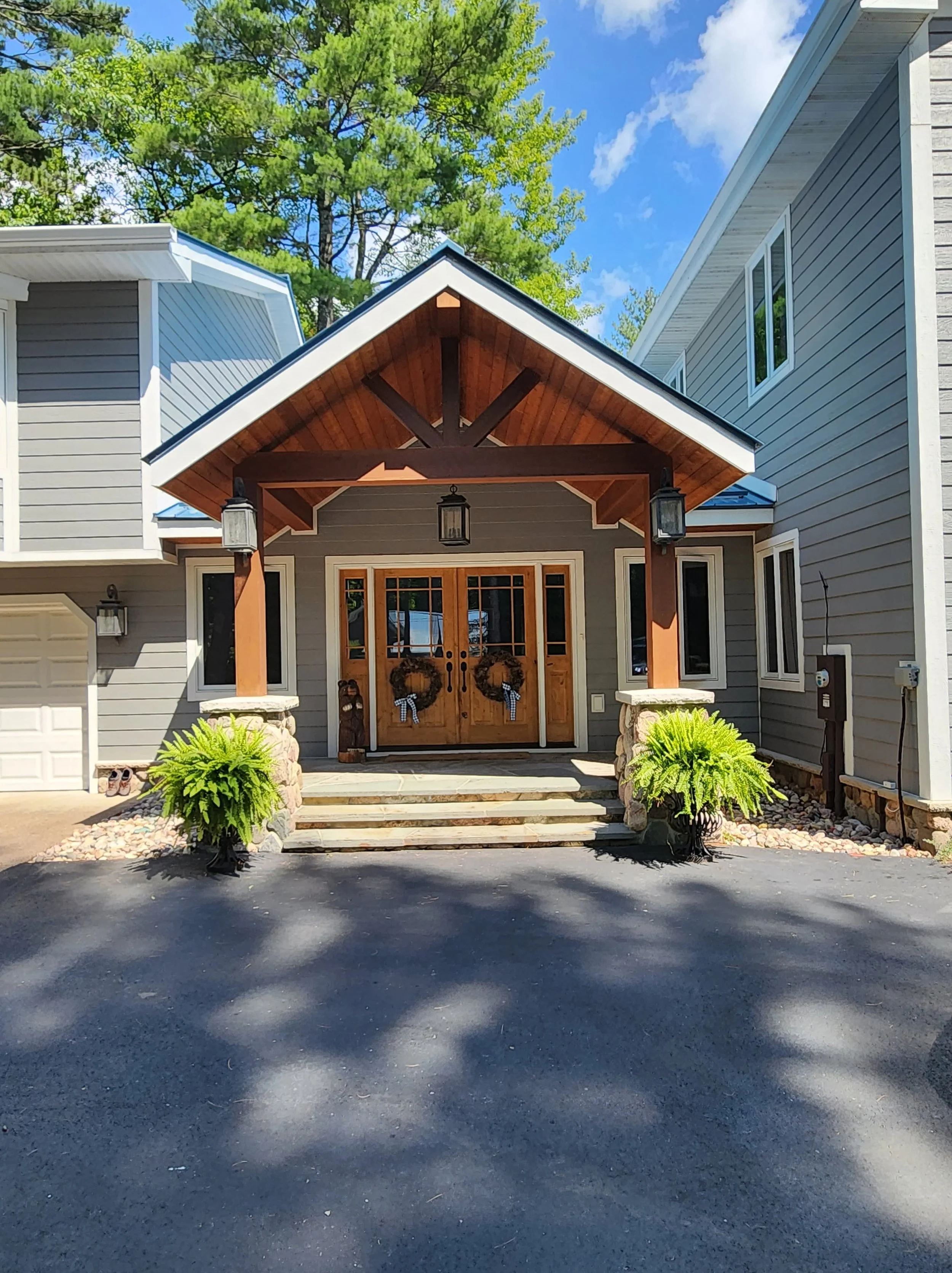 Front view of a house with a wooden front door decorated with wreaths, stone steps, potted ferns, two owl statues, and exterior lanterns, with neighboring houses and trees in the background.