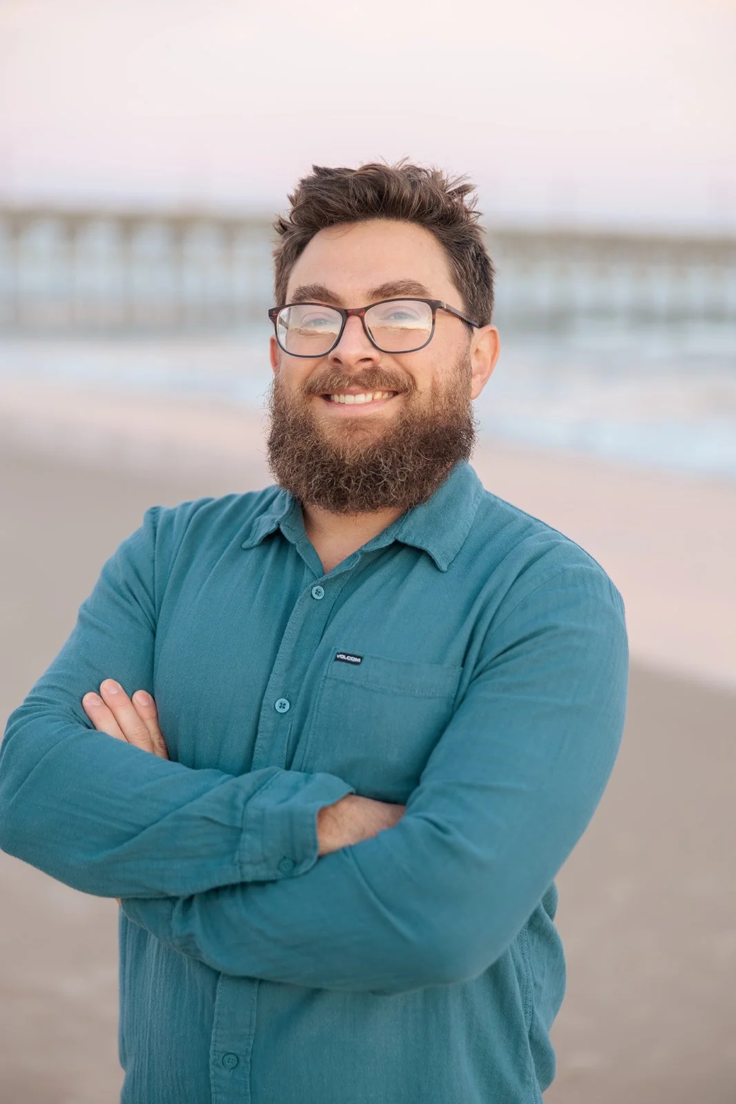 A man with glasses and a beard, smiling at the camera, standing on a beach with arms crossed, with a pier visible in the background.