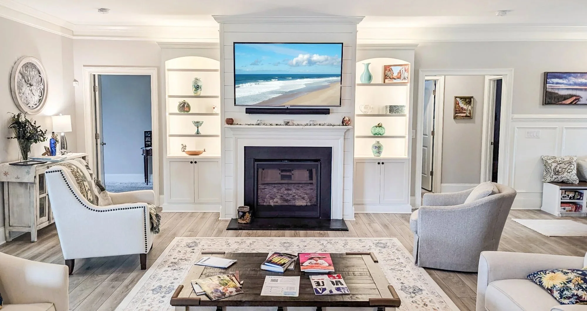 Living room with white walls, a fireplace, and a large flat-screen TV showing a beach scene. There are built-in shelves with decorative items, armchairs, a wooden coffee table with magazines, and a patterned rug.