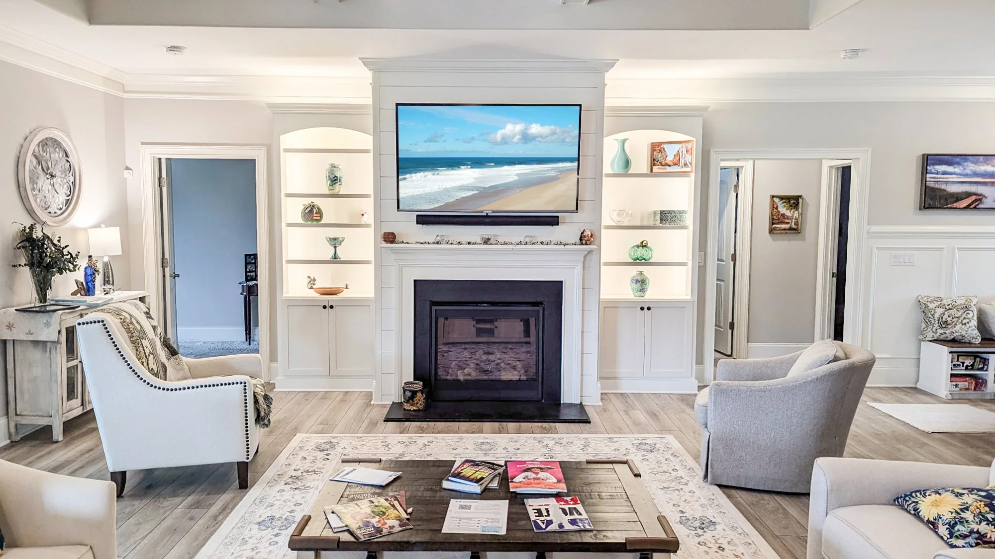 Living room with white walls, a fireplace, and built-in shelves, decorated with vases and artwork. A flat-screen TV displays a beach scene above the fireplace. There are armchairs and a coffee table with magazines and brochures.