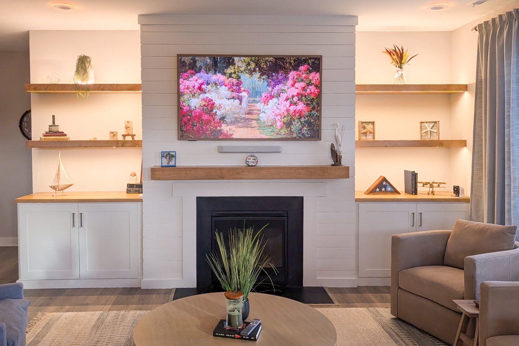 Living room with a white brick fireplace, a mounted flat-screen TV displaying a colorful garden scene, and built-in shelves with decorative items. A beige armchair and a round coffee table with a plant and remote controls are in the foreground.