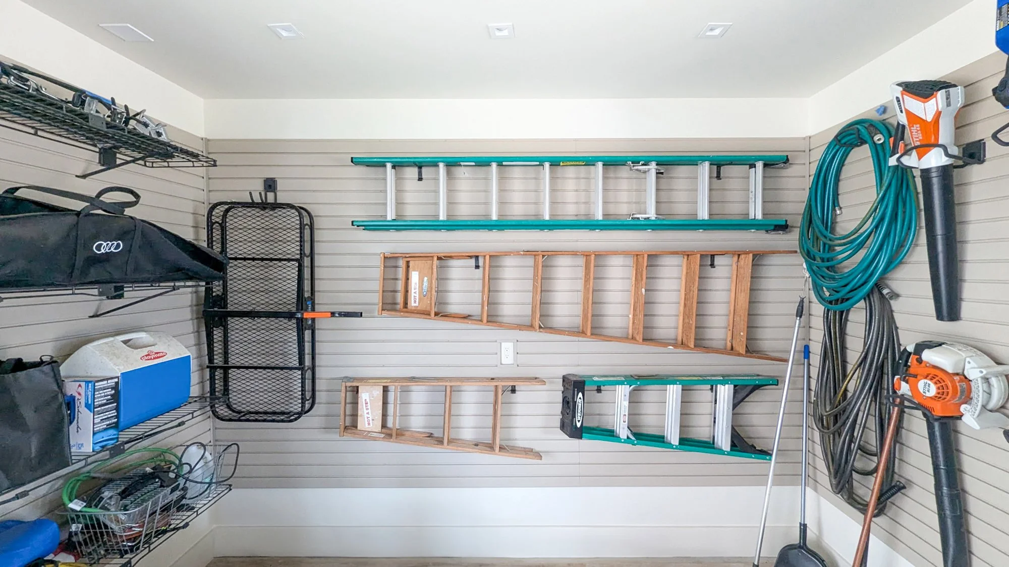 Garage wall with storage shelves holding ladders, garden hose, leaf blower, and various tools and storage bins.