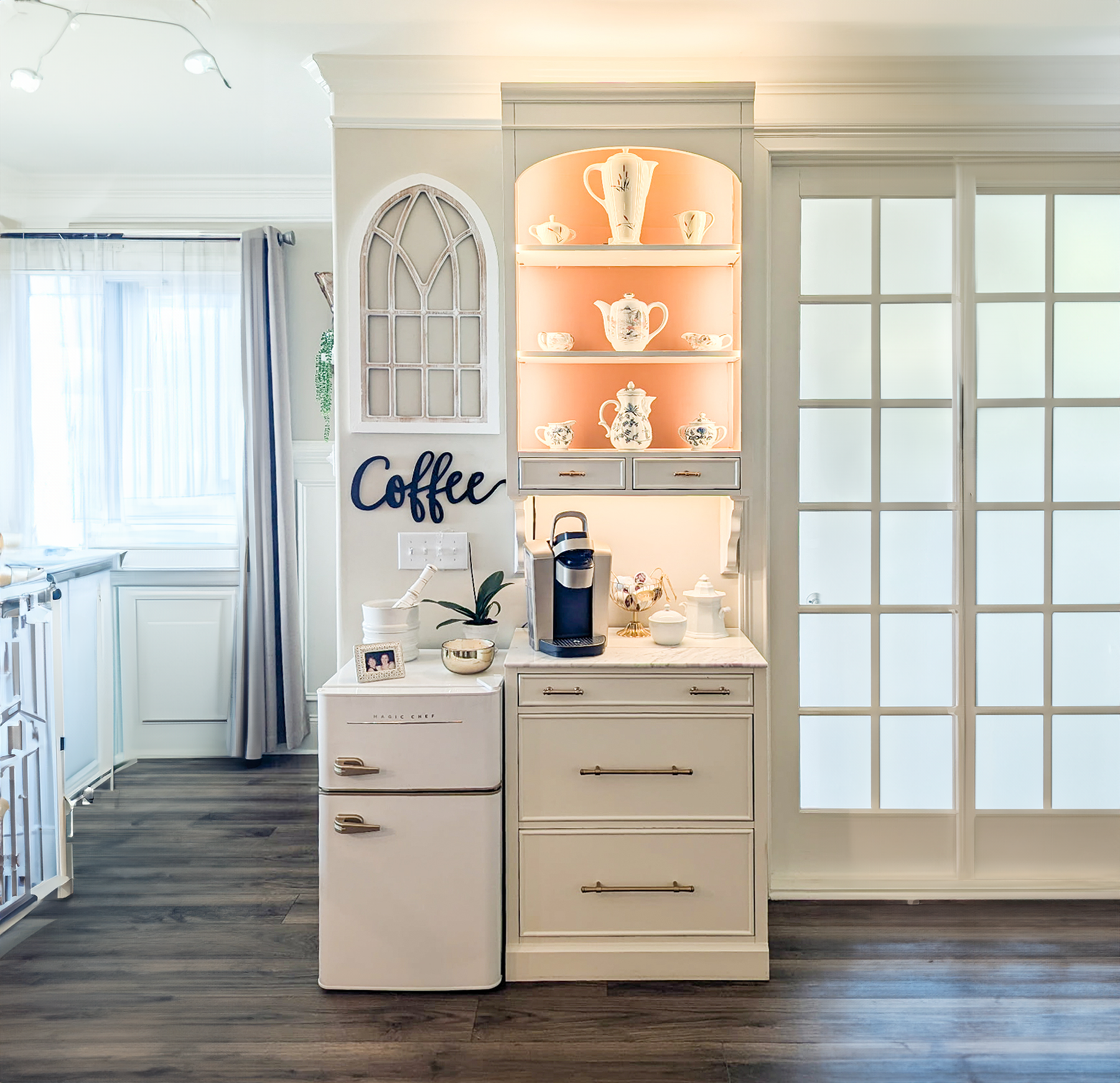 Kitchen corner with a small white fridge, a coffee machine, a decorative plant, and a display shelf with teapots and cups. The wall has a sign that says 'Coffee' and a large window with curtains nearby.