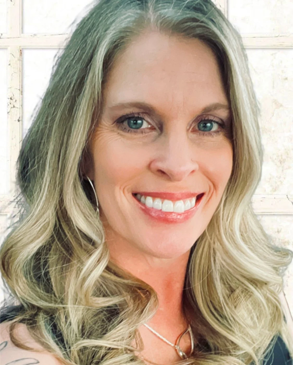 Close-up of a woman with long blonde curly hair, green eyes, and a bright smile, wearing subtle earrings and a necklace, with a light, neutral background.