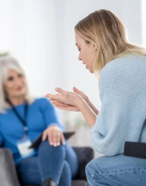 A woman with blonde hair in a blue sweater sitting and talking with another woman with blonde hair and a blue shirt in a bright, modern room.