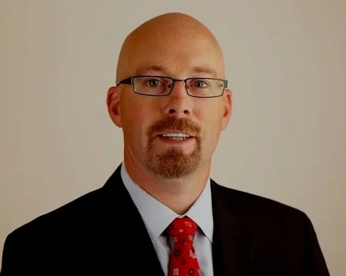 A man with glasses, a goatee, and a bald head wearing a suit, dress shirt, and red tie, posing against a plain background.
