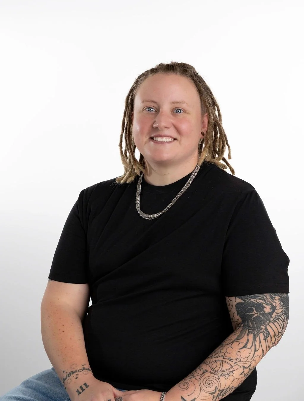 A woman with dreadlocks wearing a black shirt and silver jewelry, smiling against a white background.