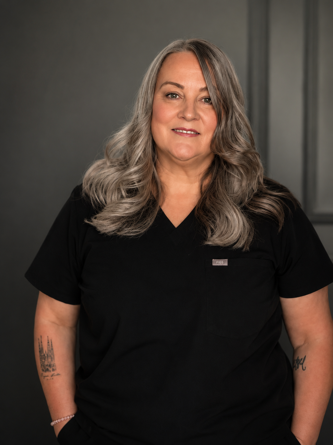 A middle-aged woman with long wavy silver hair, wearing a black medical uniform with a small logo on the chest, posing against a gray background.