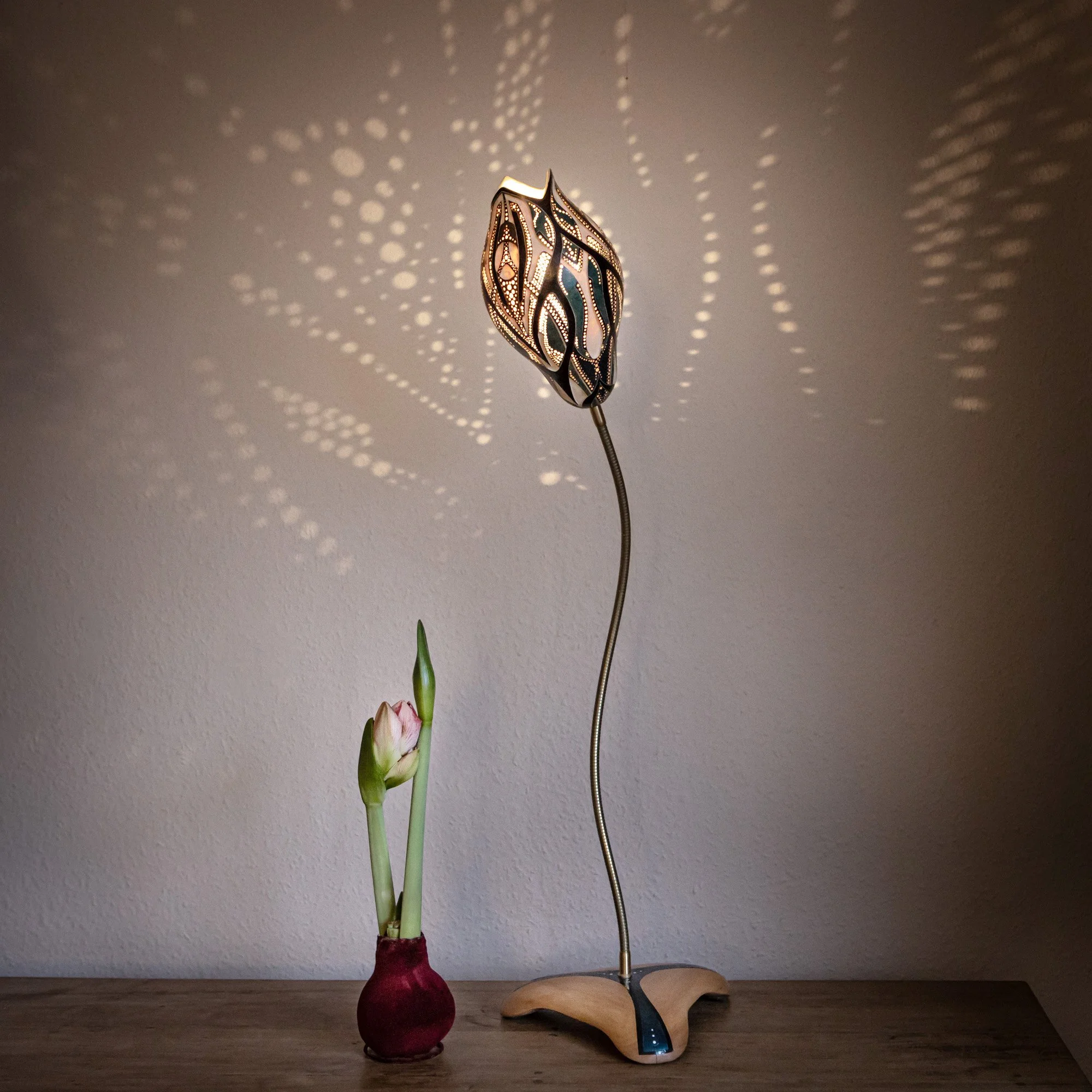 Ein stilvoller Tischlampe mit einem Muster, das komplexe Lichtmuster an die Wand projiziert, neben einer blühenden Pflanze in einer roten Zwiebel.