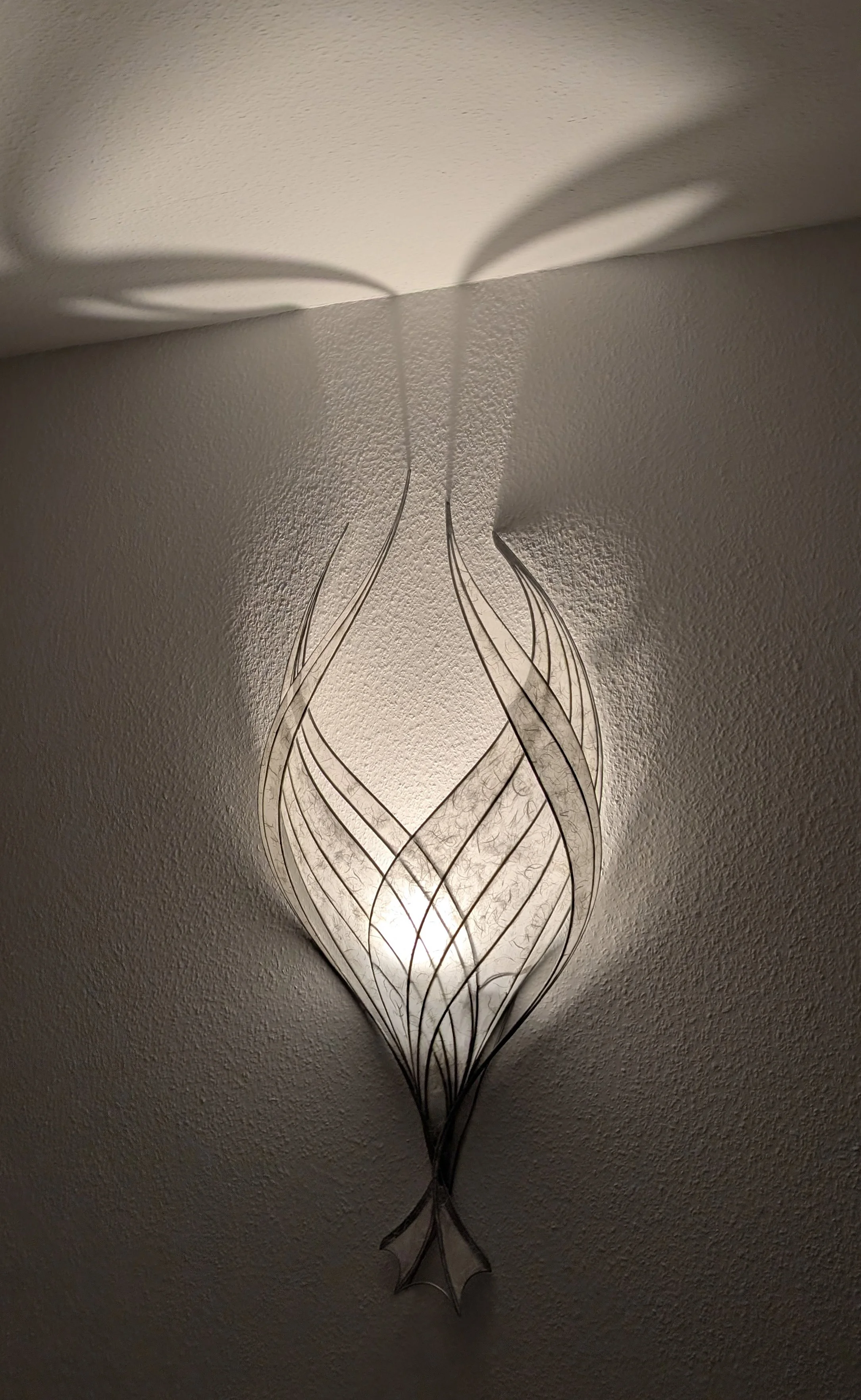 Wandleuchte mit geschwungenem, filigranem Metallrahmen, der ein Muster bildet, das an eine Blume oder einen Flasche erinnert, beleuchtet an einer Zimmerdecke