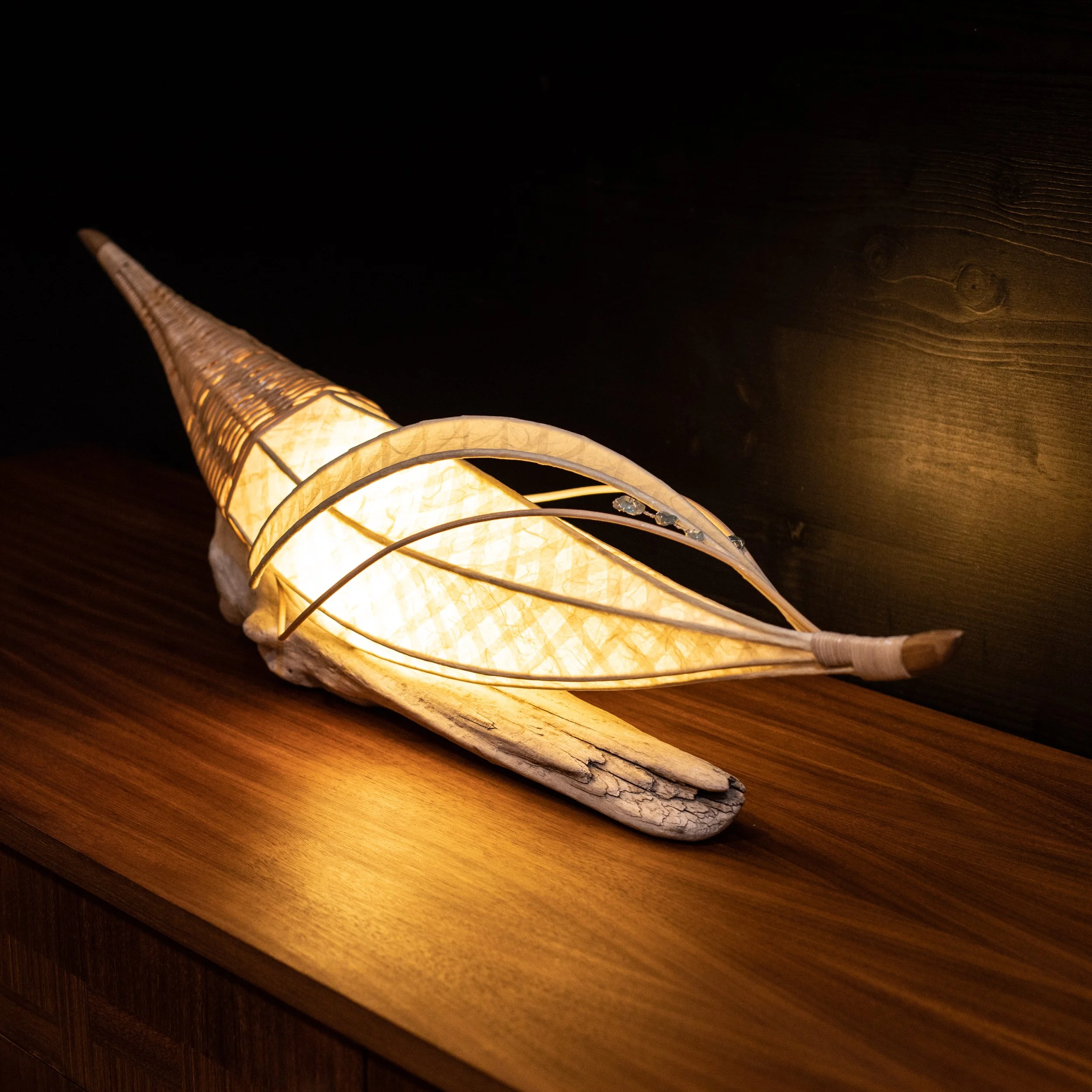 Eine dekorative Lampe in Form eines spiralförmigen Horns, das aus Holz und dünnen Metallstreifen besteht, auf einem Holzoberfläche.