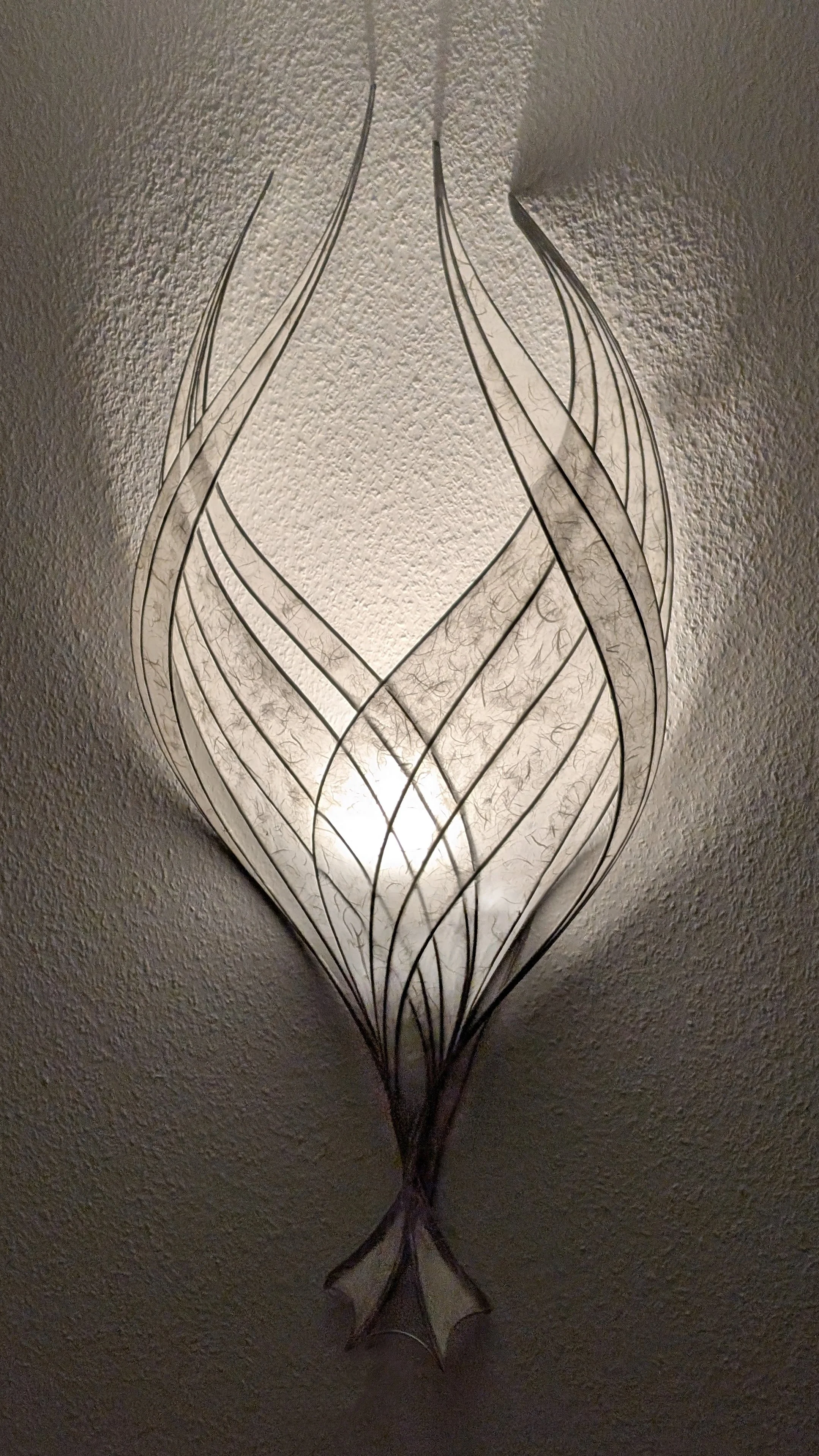 Hängelampe mit filigraner, geschwungener Metallstruktur, die ein elegantes Muster bildet, beleuchtet die Decke und die Wand dahinter.