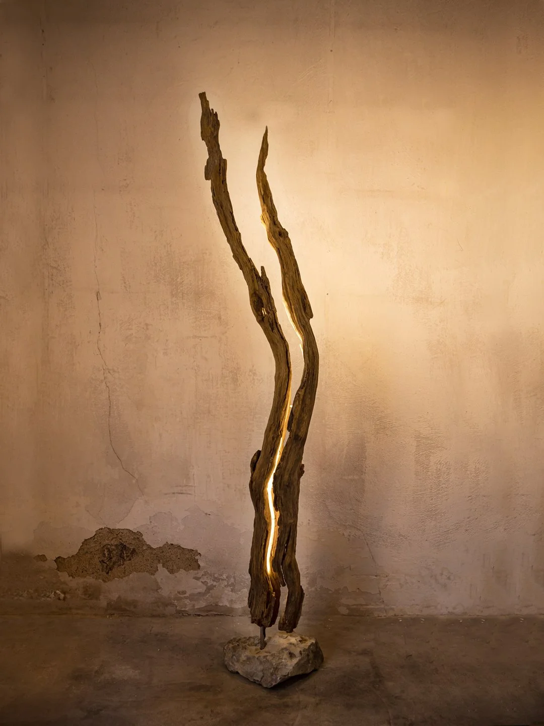 Skulptur aus zwei langen, unregelmäßig geformten Holzstücken, die an einem Stein befestigt sind und eine vertikale Linie mit einer eingebauten Beleuchtung bilden, vor einer uneben verputzten Wand.