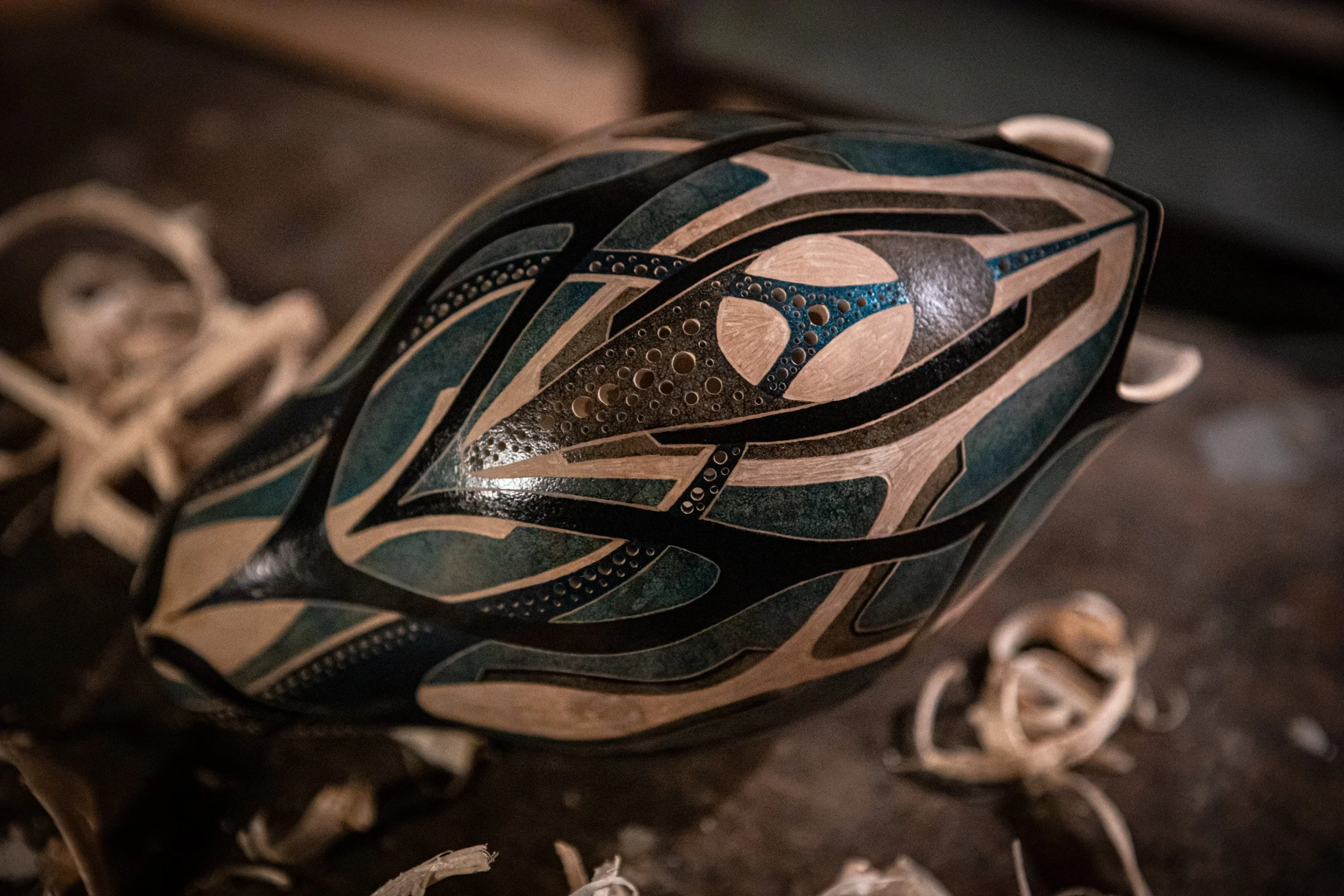 Ein handgefertigtes Holzkunstwerk mit einem komplexen, stilisierten Vogelmotiv in Schwarz, Blau, Beige und Grau, das auf einem dunklen Untergrund liegt, umgeben von Holzspänen.