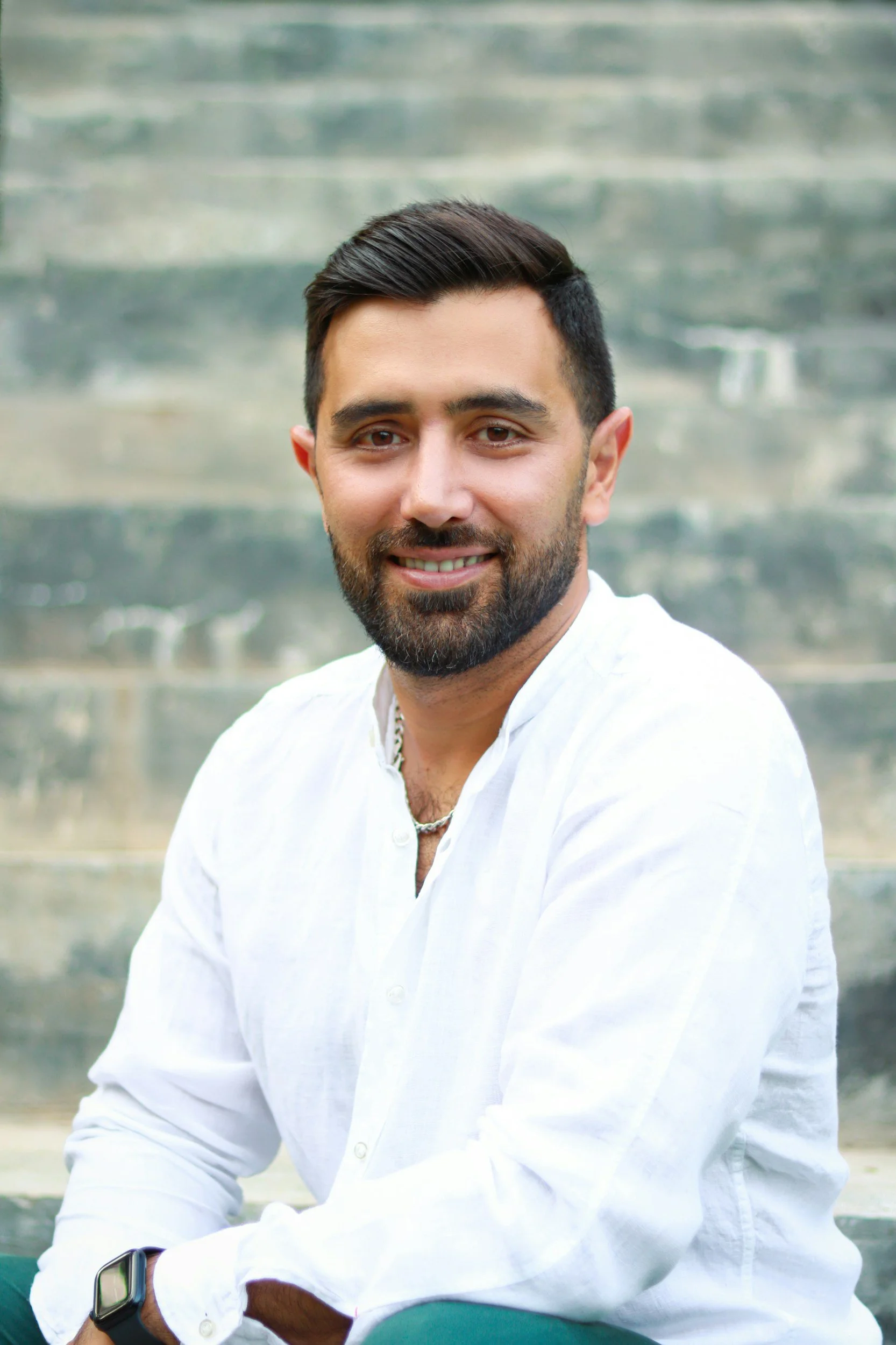 A smiling man with dark hair and beard, wearing a white shirt, sitting outdoors against a blurred stone staircase background.