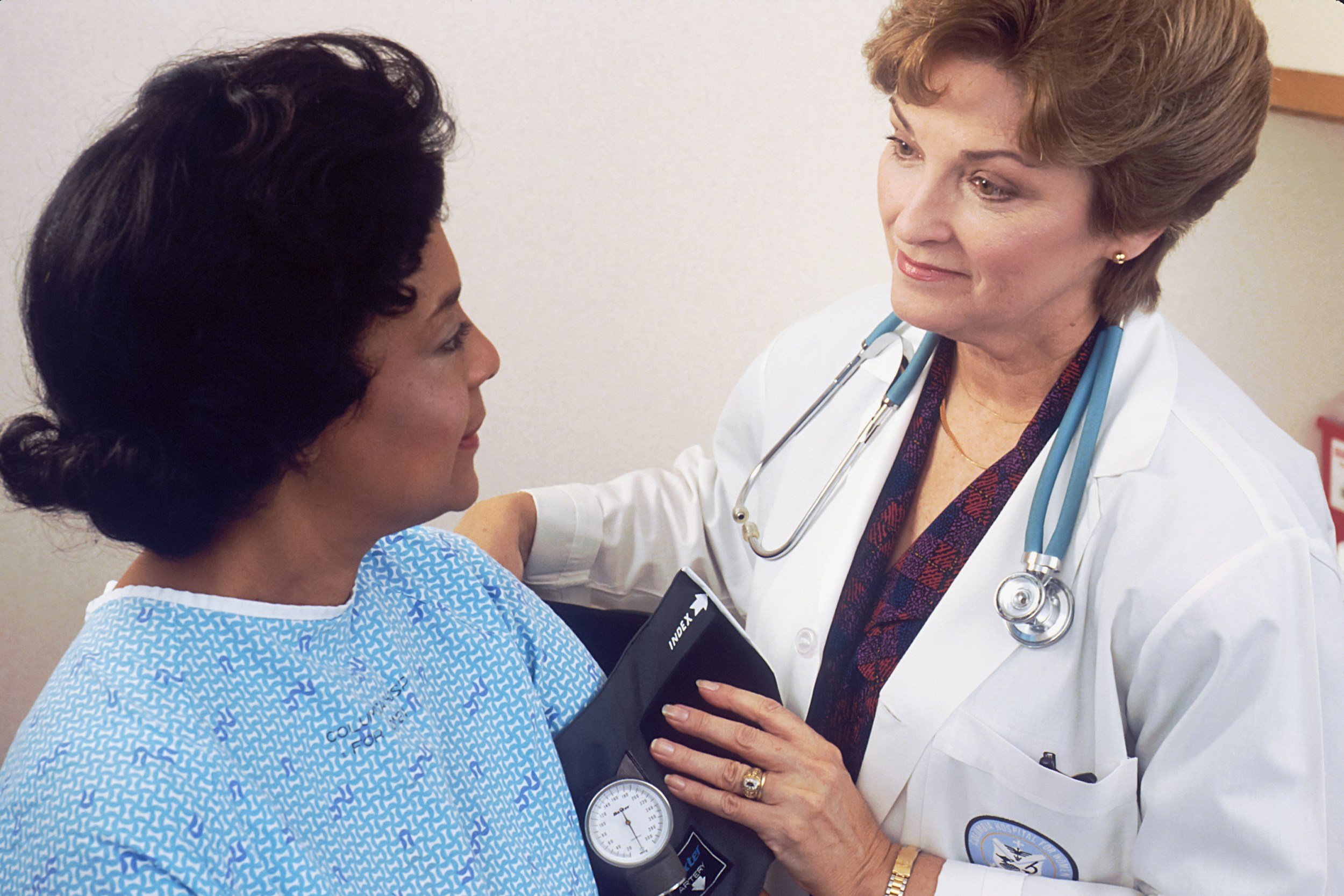 A female doctor with short, light brown hair and a stethoscope around her neck is examining and speaking to a female patient with dark hair wearing a hospital gown. The doctor is holding a blood pressure cuff and the patient is sitting on an examination table.
