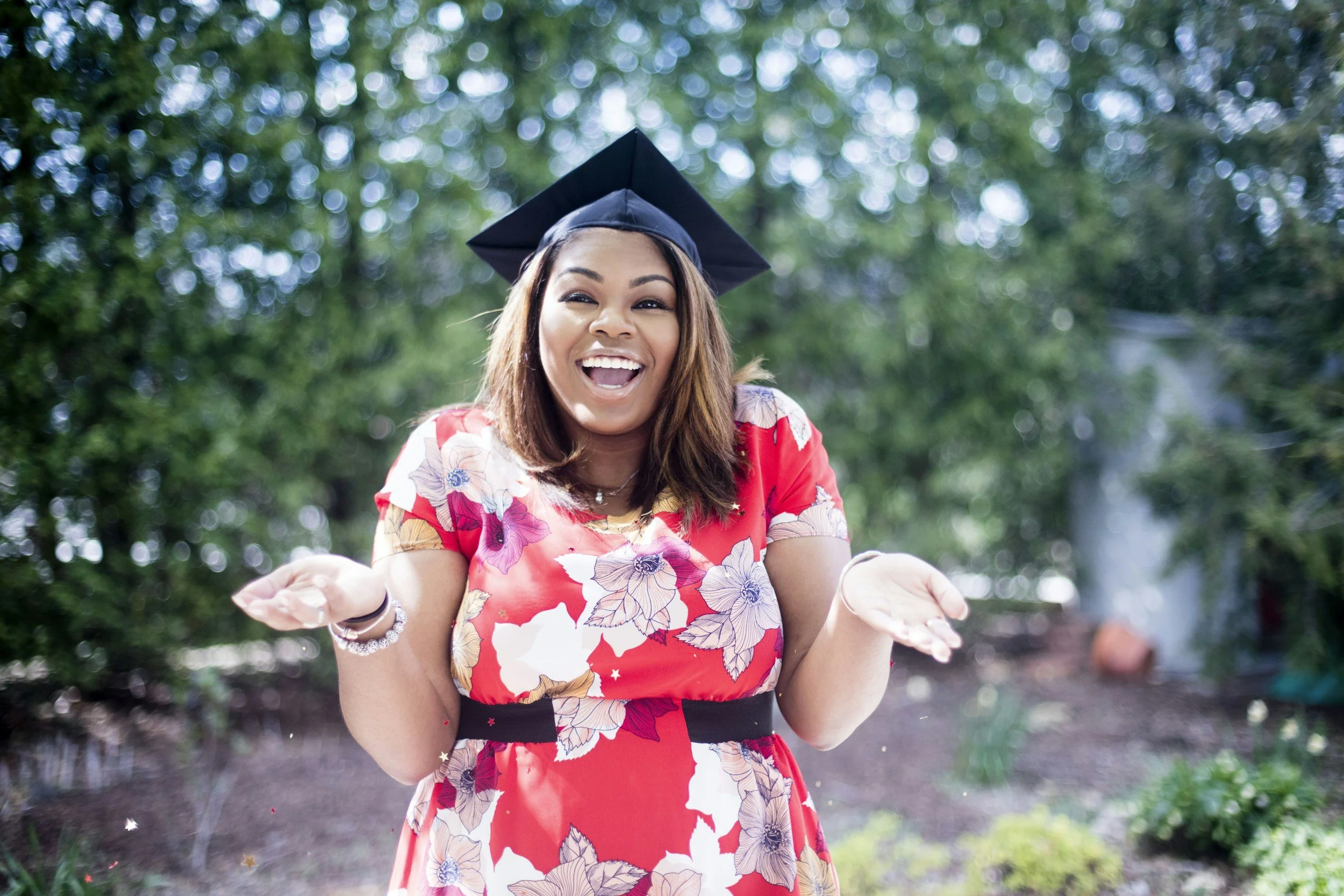 Smiling woman wearing a red floral dress and a graduation cap outdoors with trees in the background, celebrating graduation.
