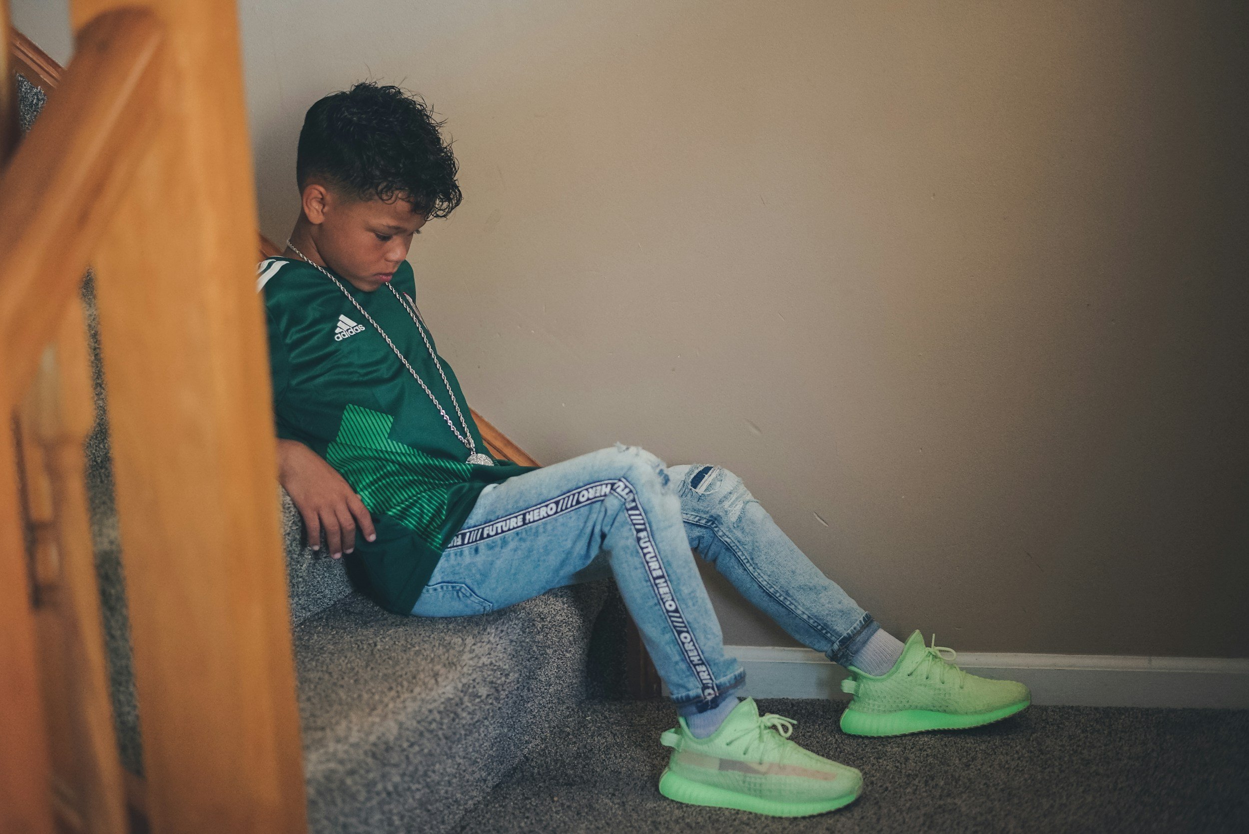 A young boy sitting on carpeted stairs with a beige wall behind him, wearing a green sports jersey, ripped jeans, neon green sneakers, and a silver chain.