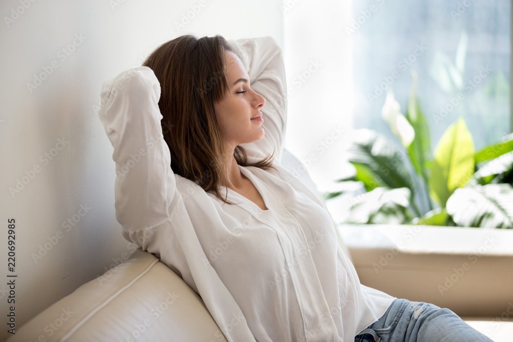 A woman relaxing on a cream sofa with her eyes closed and arms behind her head, sitting near a window with sunlight and greenery outside.